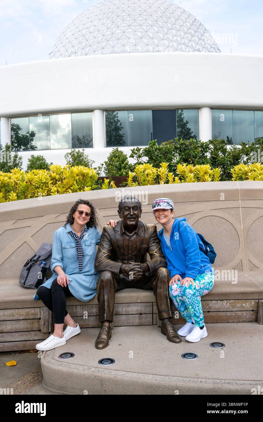 Glückliche Besucher lächeln für die Kamera, während sie neben einer Bronzestatue von Walt Disney am Eingang von EPCOT sitzen. Stockfoto