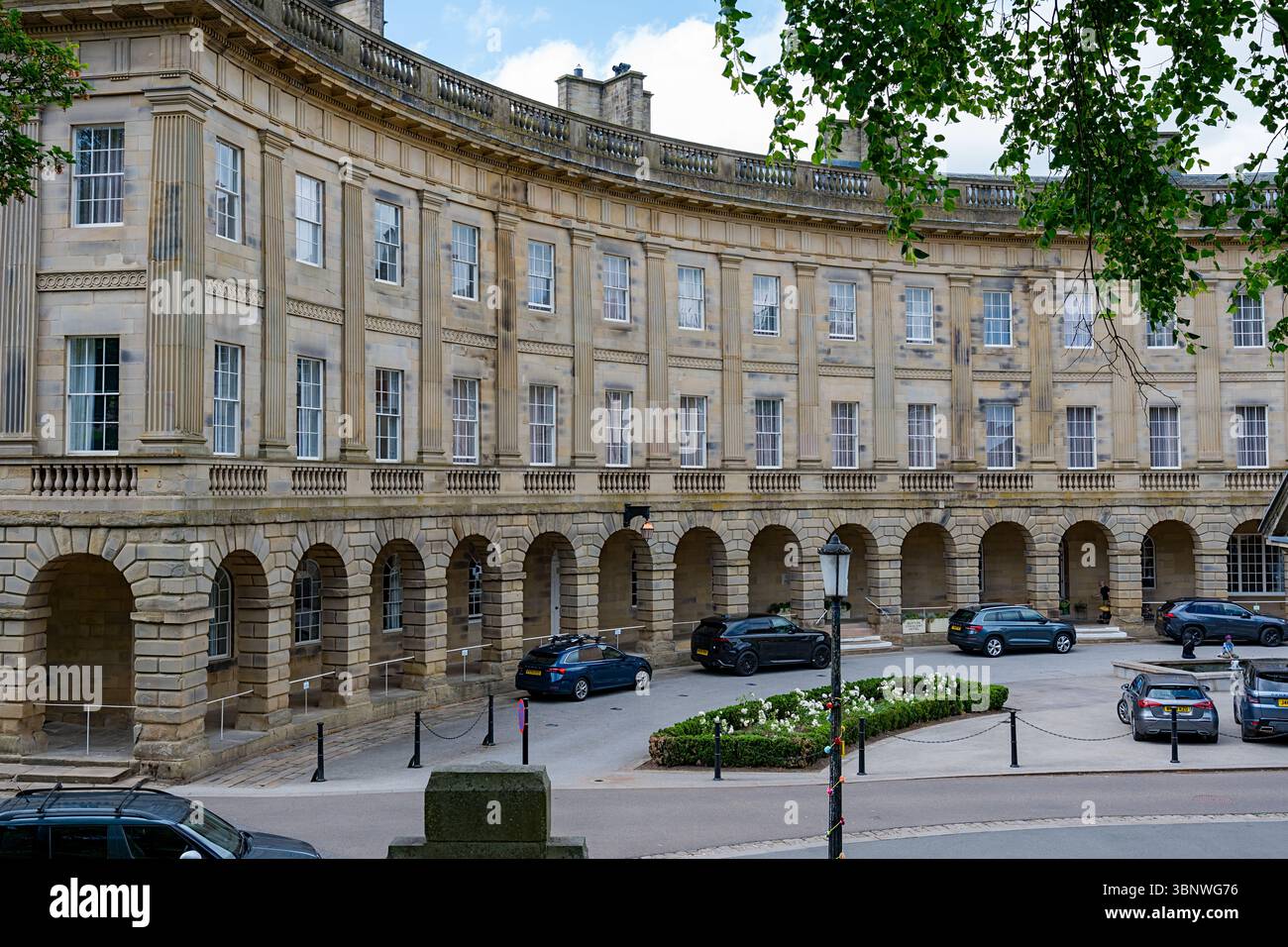 Buxton, Derbyshire, Großbritannien, 28. Juni 2025: Historisches Halbmondgebäude Buxton mit geparkten Autos und üppigem Grün. Stockfoto