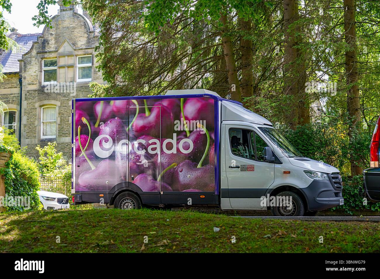 Buxton, Derbyshire, Großbritannien, 28. Juni 2025: Ocado Food Delivery Truck mit lebendigem Kirschdesign in einer üppigen, grünen Wohngegend. Stockfoto