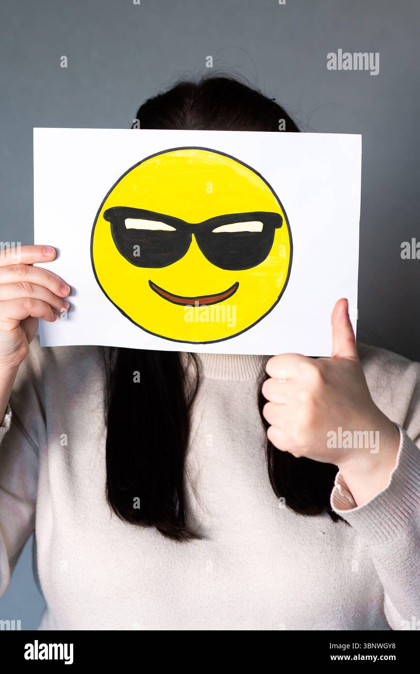 Frau, die ein glückliches Emoji mit Sonnenbrillen-Maske vor dem Gesicht hält. Daumen nach oben. Welt-Emoji-Tag 17. Juli. Stockfoto
