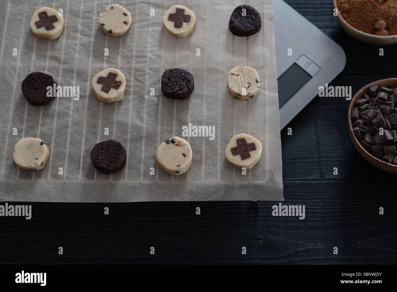 Vorderansicht des Keksteigs auf Backpapier mit Schüssel mit Schokoladenchips, brauner Zuckerschale und digitaler Skala auf schwarzer Holzoberfläche Stockfoto