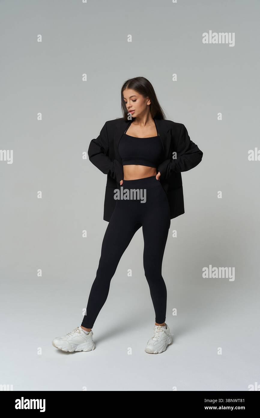 Selbstbewusste Frau in dunkler Sportkleidung und Turnschuhen posiert mit Händen auf der Hüfte. Sie trägt eine schwarze Jacke über einem passenden Crop-Top und Leggings. Stockfoto