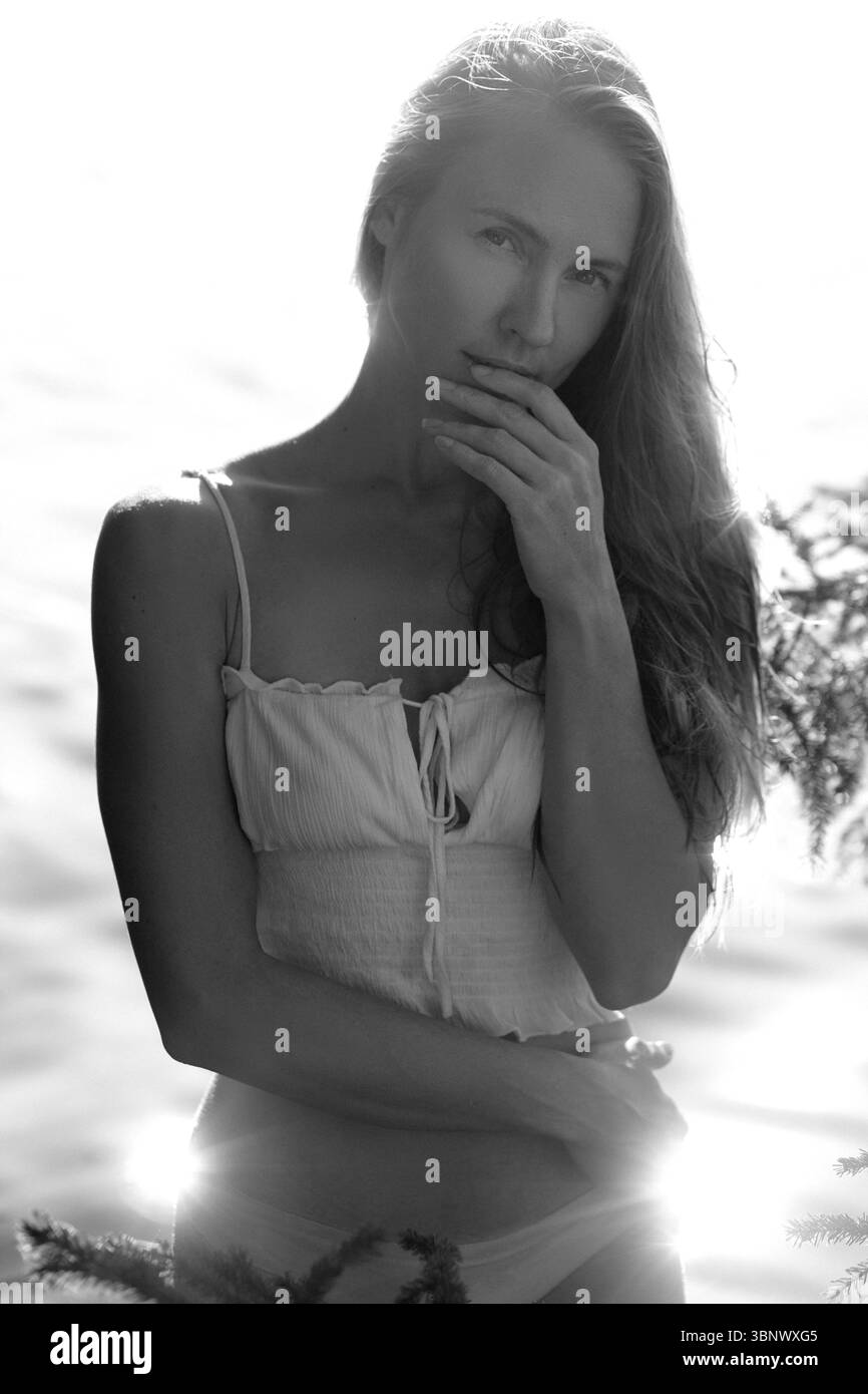 Die Frau steht nahe am Wasser und berührt sanft ihr Gesicht mit einem besinnlichen Ausdruck. Das Sonnenlicht erzeugt ein sanftes Leuchten um sie herum, das die Ruhe verstärkt Stockfoto