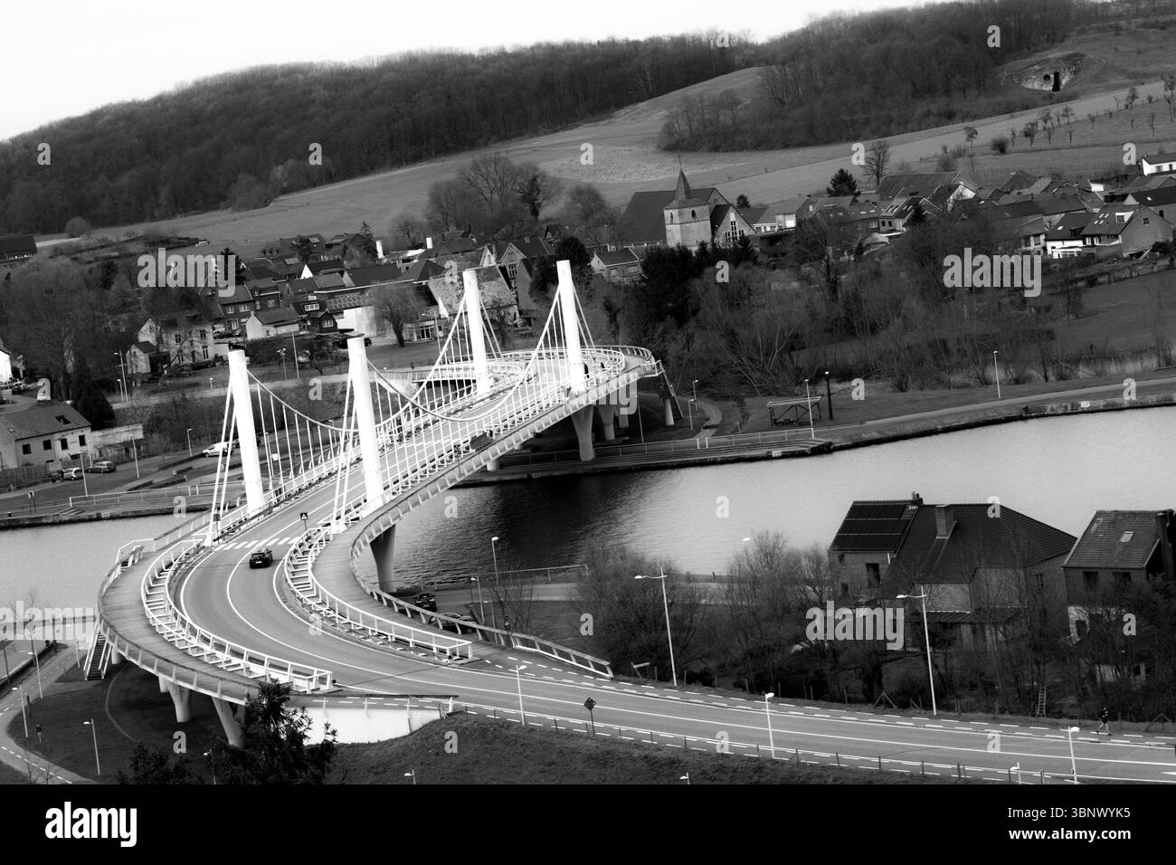 Eine geschwungene moderne Hängebrücke in Schwarz-weiß, die einen Fluss mit einem ländlichen Dorf und bewaldeten Hügeln im Hintergrund an einem bewölkten Tag überspannt. Stockfoto