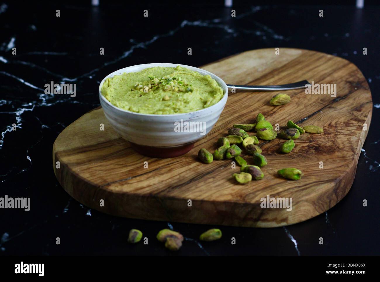Pistaziencreme in handgemachter Schüssel mit verstreuten Nüssen auf hölzernem Schneidebrett – dunkler Hintergrund Stockfoto