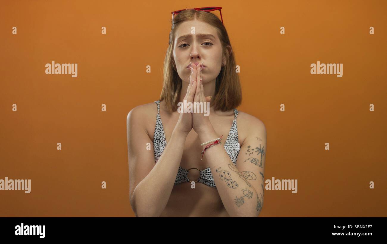 Frau mit Tätowierungen und betender Geste in einem Bikini vor einem isolierten orangen Hintergrund, die Emotionen mit roter Haarspange und Sonnenbrille ausdrückt. Stockfoto