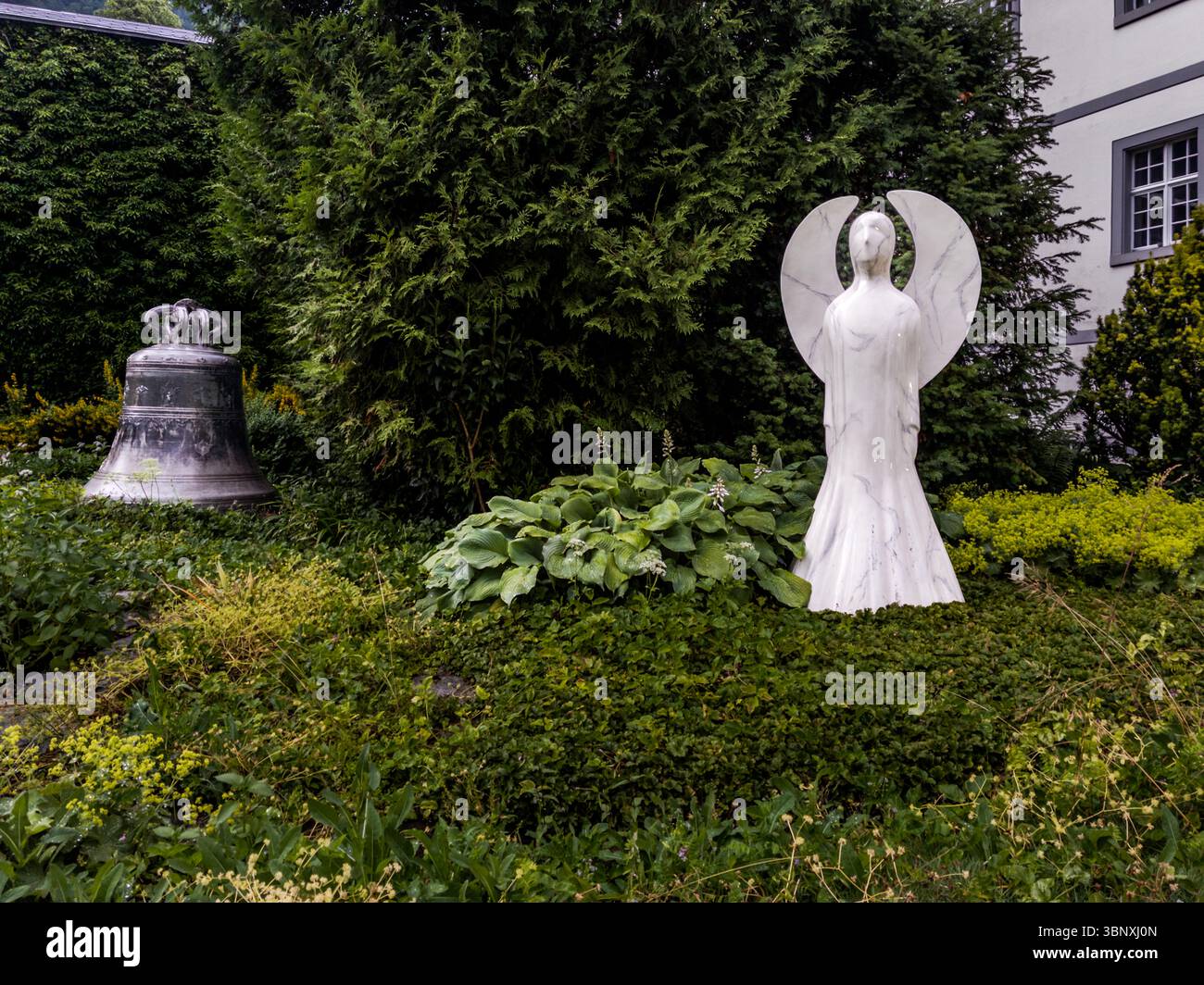 Eine historische Kirchenglocke und eine moderne Engelsstatue aus weißem Marmor stehen im Garten der Abtei Engelberg, gegründet 1120 von Konrad von Sellenbüren und eine der ältesten Benediktinerabteien der Schweiz. Am Aeschiweg in Engelberg, Kanton Obwalden, Schweiz, verbindet die Szene klösterliche Tradition mit zeitgenössischer religiöser Kunst. Stockfoto
