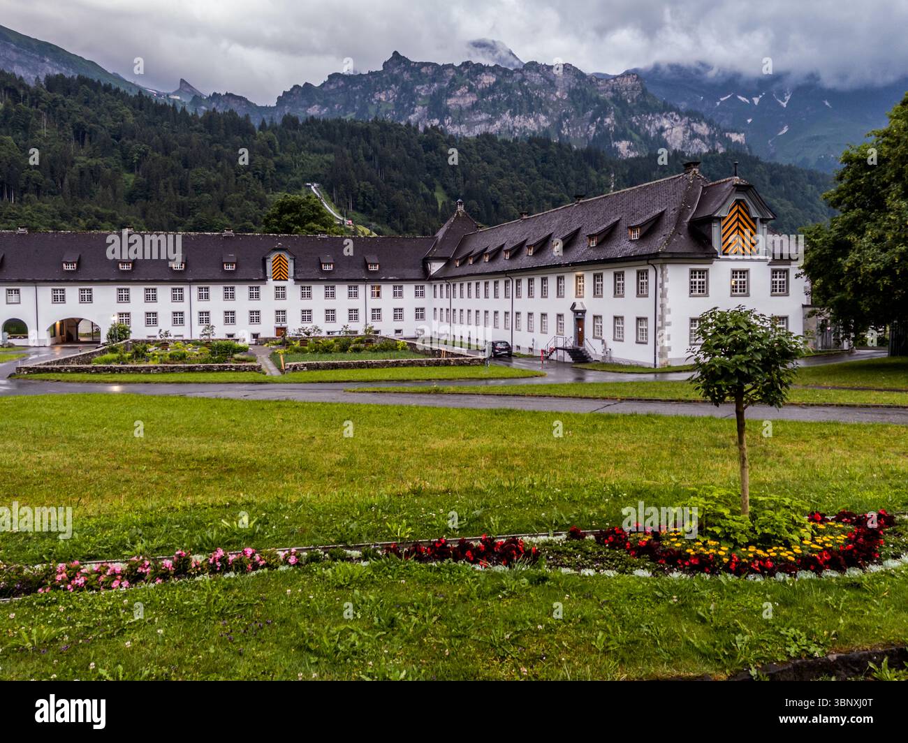Das Kloster Engelberg wurde 1120 von Konrad von Sellenbüren gegründet und ist eine der ältesten Benediktinerabteien der Schweiz. Die Klosteranlage ist eines der bedeutendsten Barockensembles der Zentralschweiz und verfügt über die größte Orgel des Landes. Das Benediktinerkloster Engelberg, ein markantes weißes Gebäude mit dunklem Dach, ist von grünen Rasenflächen und bunten Blumenbeeten umgeben. Die majestätischen Berge und eine Schanze sind im Hintergrund zu sehen. Aeschiweg, Engelberg, Obwalden, Schweiz Stockfoto