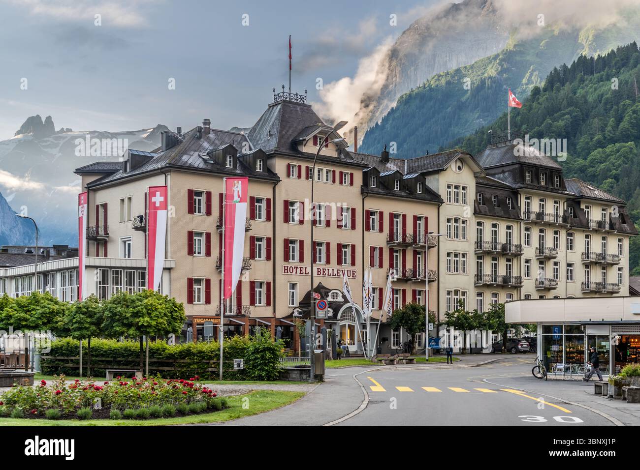 Das Hotel Bellevue in Engelberg, Schweiz, präsentiert sich vor der beeindruckenden Kulisse der Schweizer Alpen. Die Schweizer Flagge und die Flagge des Kantons Obwalden fliegen vor dem historischen Gebäude, während sich Wolken um die majestätischen Berggipfel sammeln. Die Szene fängt die malerische Schönheit der alpinen Landschaft und die traditionelle Architektur des Ortes ein Stockfoto