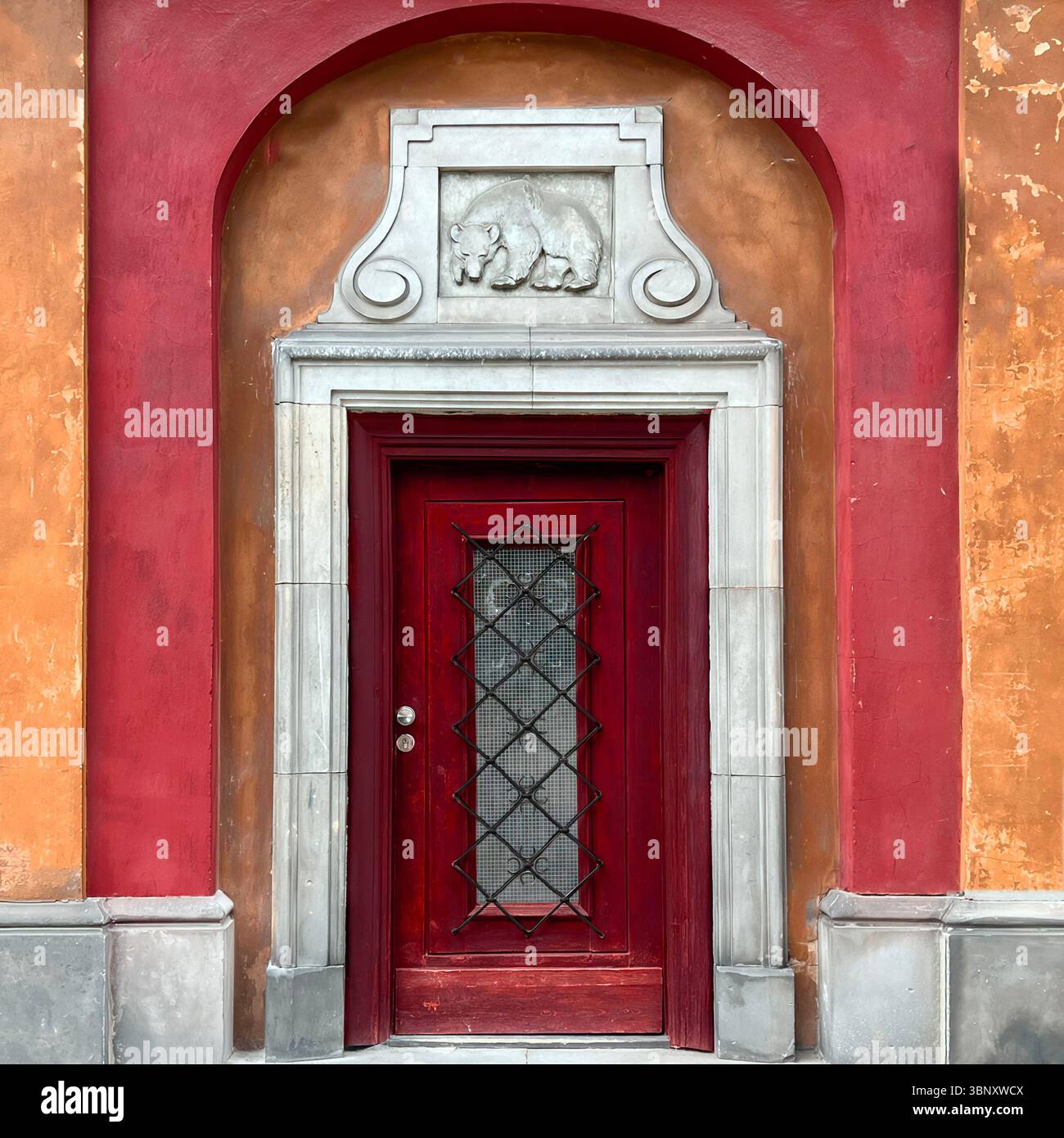 Eine tiefrote Tür in der Warschauer Altstadt steht unter einem Bärenrelief und einem Steinrahmen – ein intimes Portal voller Farben, Texturen und Symbolismus. Stockfoto