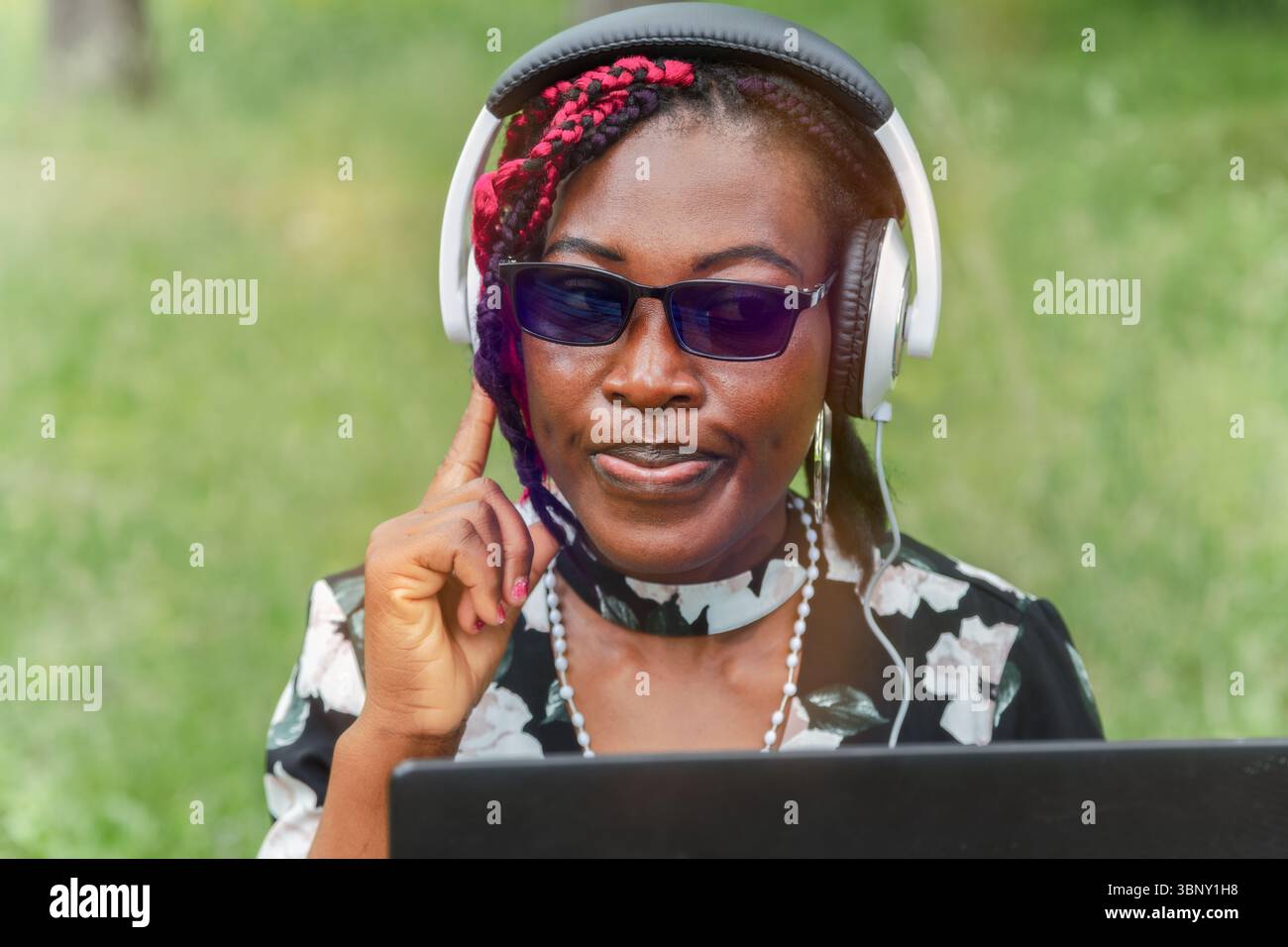Selbstbewusste junge Schwarze Frau mit Brille, die Musik mit Kopfhörern im Freien hört, während sie mit dem Laptop unterwegs ist. Perfekt für Audio-Streaming, Podcasts und Lifesty Stockfoto