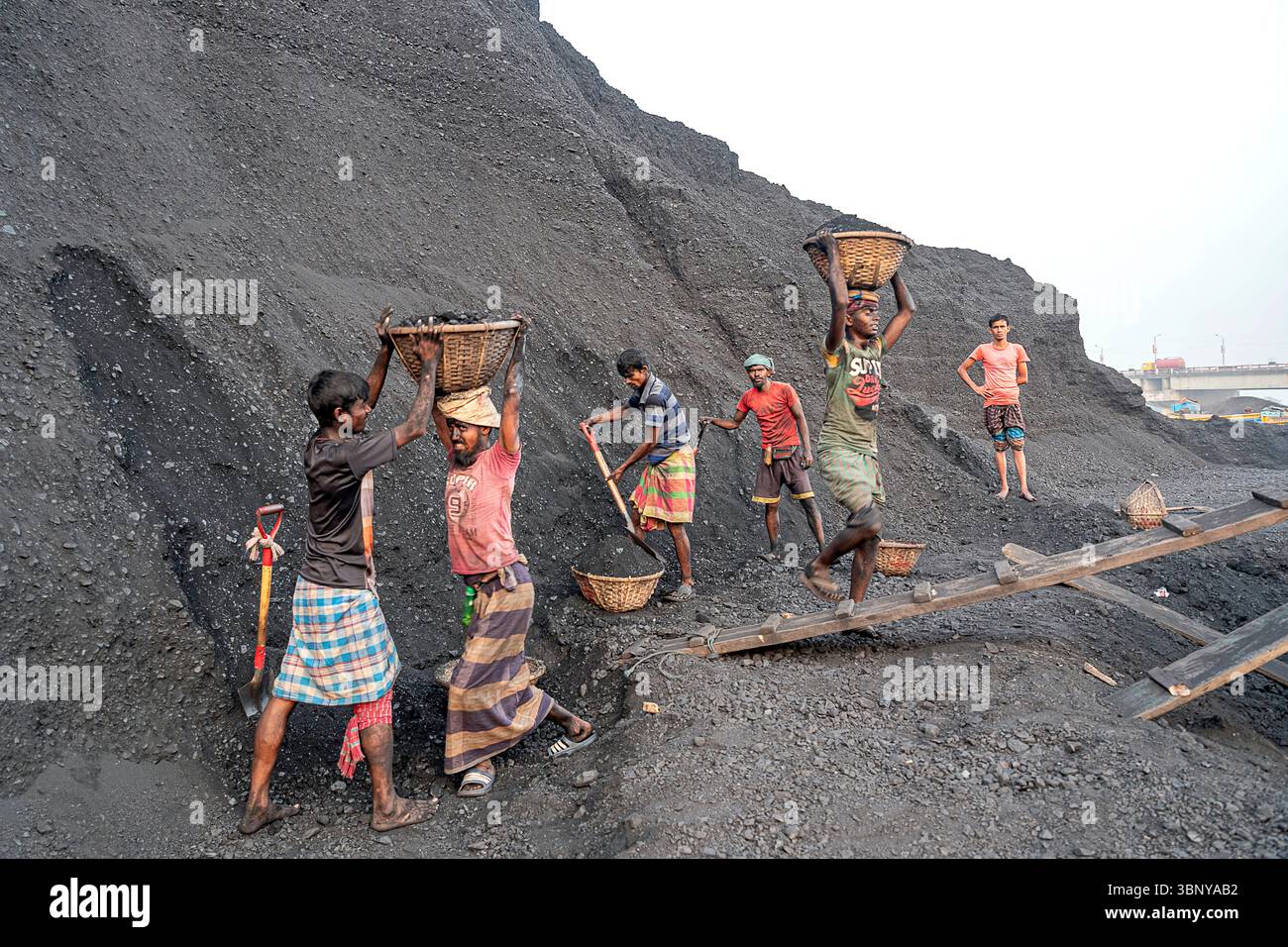 Lokale Arbeiter transportieren schwere Körbe mit Kohle an einem geschäftigen Bergbaustandort in Bangladesch bergauf und demonstrieren intensive Handarbeit und Teamarbeit. Stockfoto