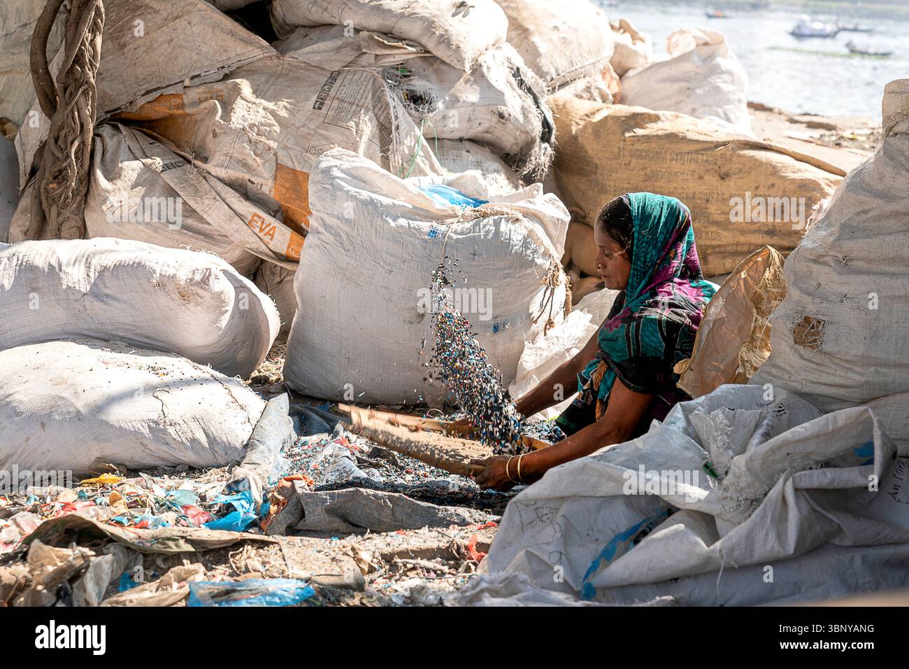 Eine Frau sortiert einen großen Stapel gemischter Plastikabfälle von Hand an einem Ufer in Bangladesch, mit Säcken gesammelter Materialien in der Nähe. Stockfoto