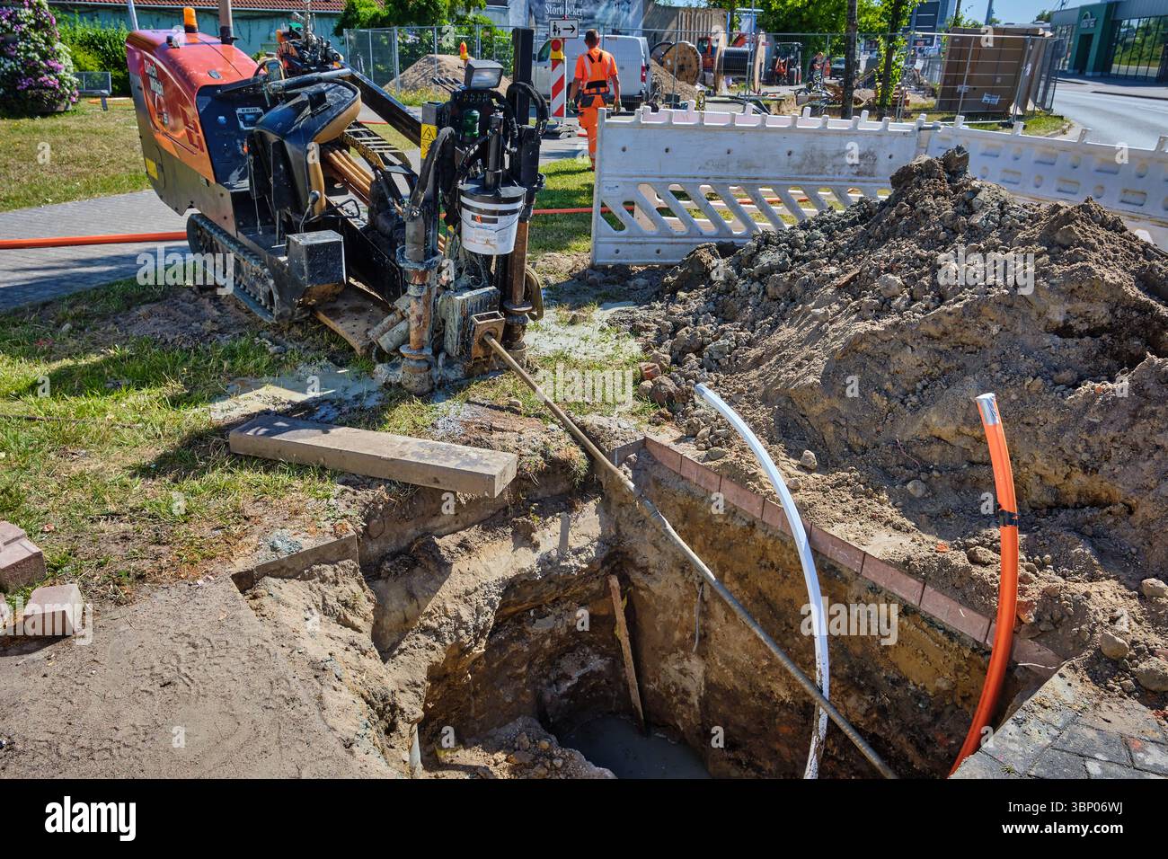 1. Juli 2025 - Stralsund-Deutschland: Kleine horizontale Bohrmaschine mit Bohrer von Startgrube an Kreisverkehrecke, orange-weiße Kabel, Sandhaufen, Stockfoto