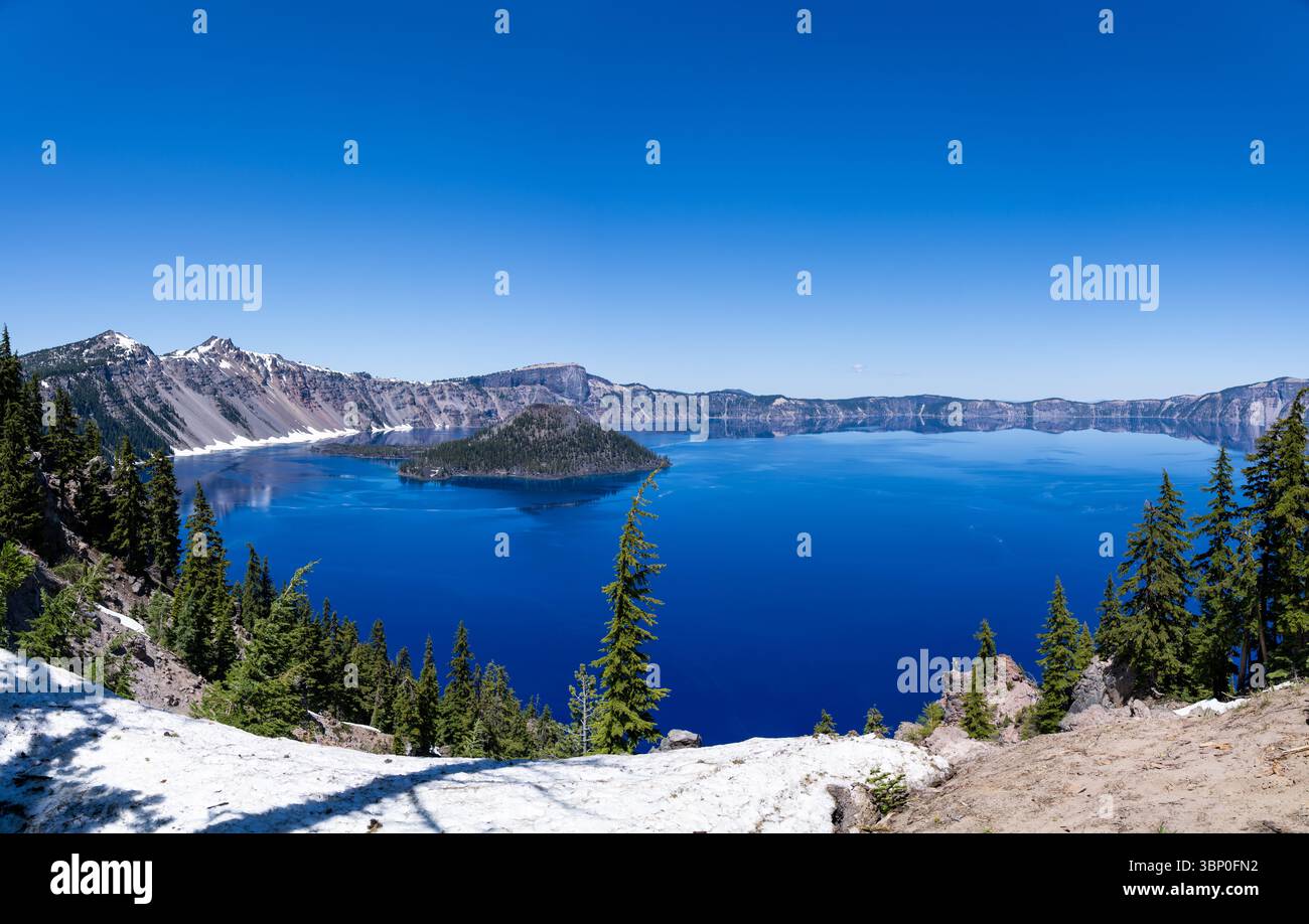 Panoramablick auf das leuchtend blaue Wasser des Crater Lake und Wizard Island, umgeben von steilen vulkanischen Kalderawänden unter einem klaren Himmel, Oregon Stockfoto