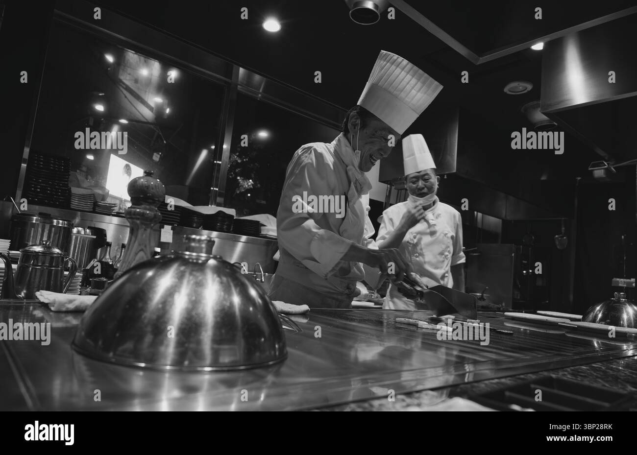 Zwei lächelnde Teppanyaki-Köche bereiten Speisen auf dem Grill zu und sorgen so für ein warmes und einladendes kulinarisches Erlebnis Stockfoto