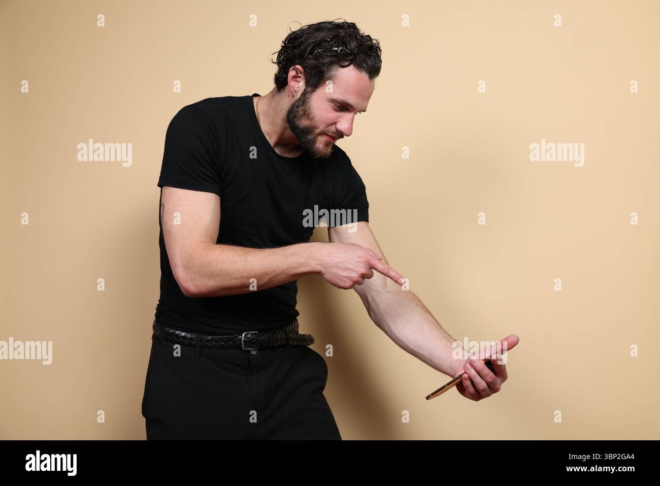 Ein fokussierter Mann hält ein Smartphone und zeigt auf seinen Unterarm, möglicherweise erklärt er eine Gesundheitsmetrik oder Fitnessdaten vor einem schlichten beigen Studio-Backgrou Stockfoto