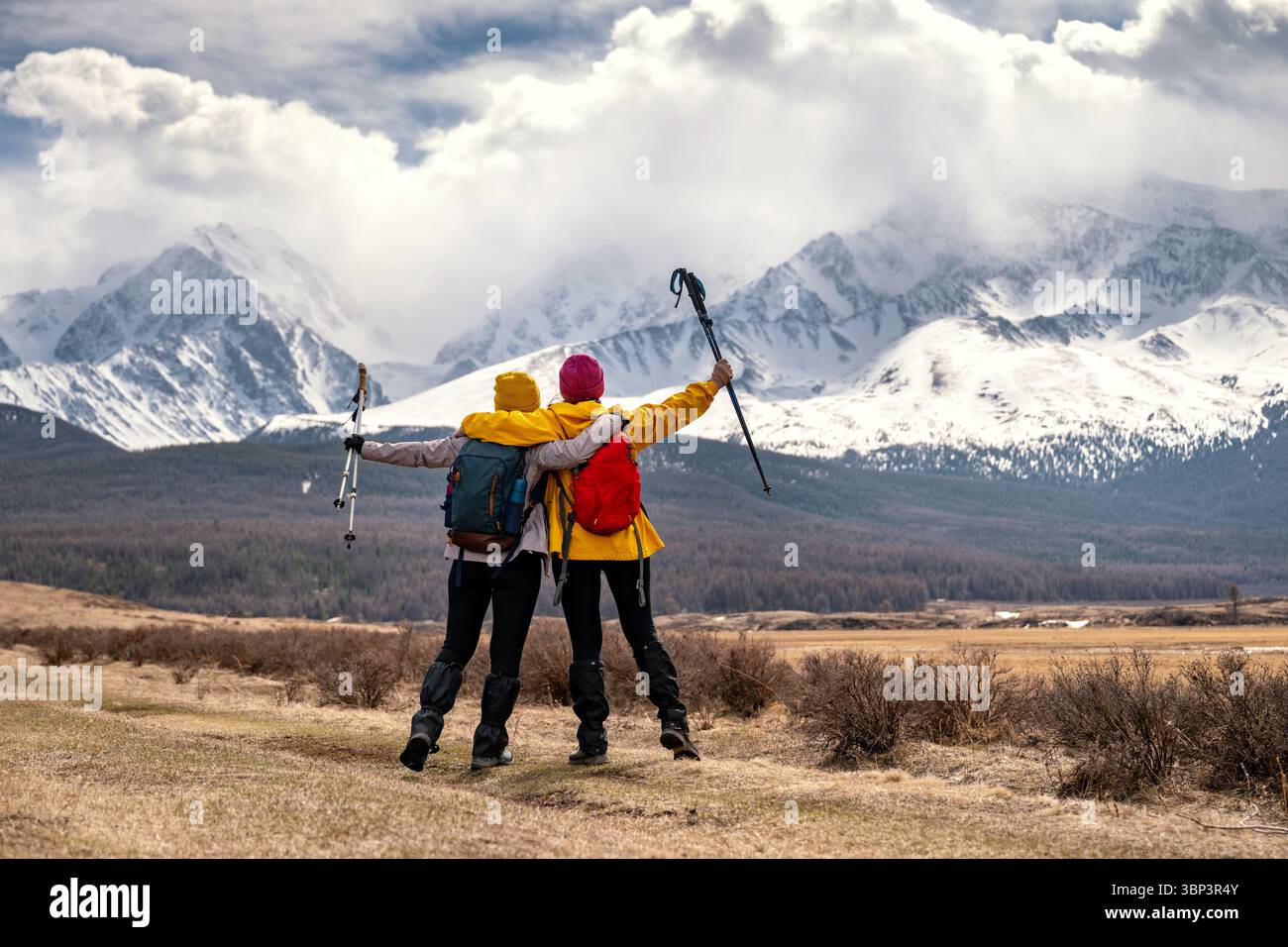 Zwei Wandererinnen mit Rucksäcken stehen umarmt und heben ihre Arme mit Trekkingstöcken an den Seiten vor dem Hintergrund der hohen schneebedeckten Gipfel im Altai-Gebirge, Blick von hinten Stockfoto
