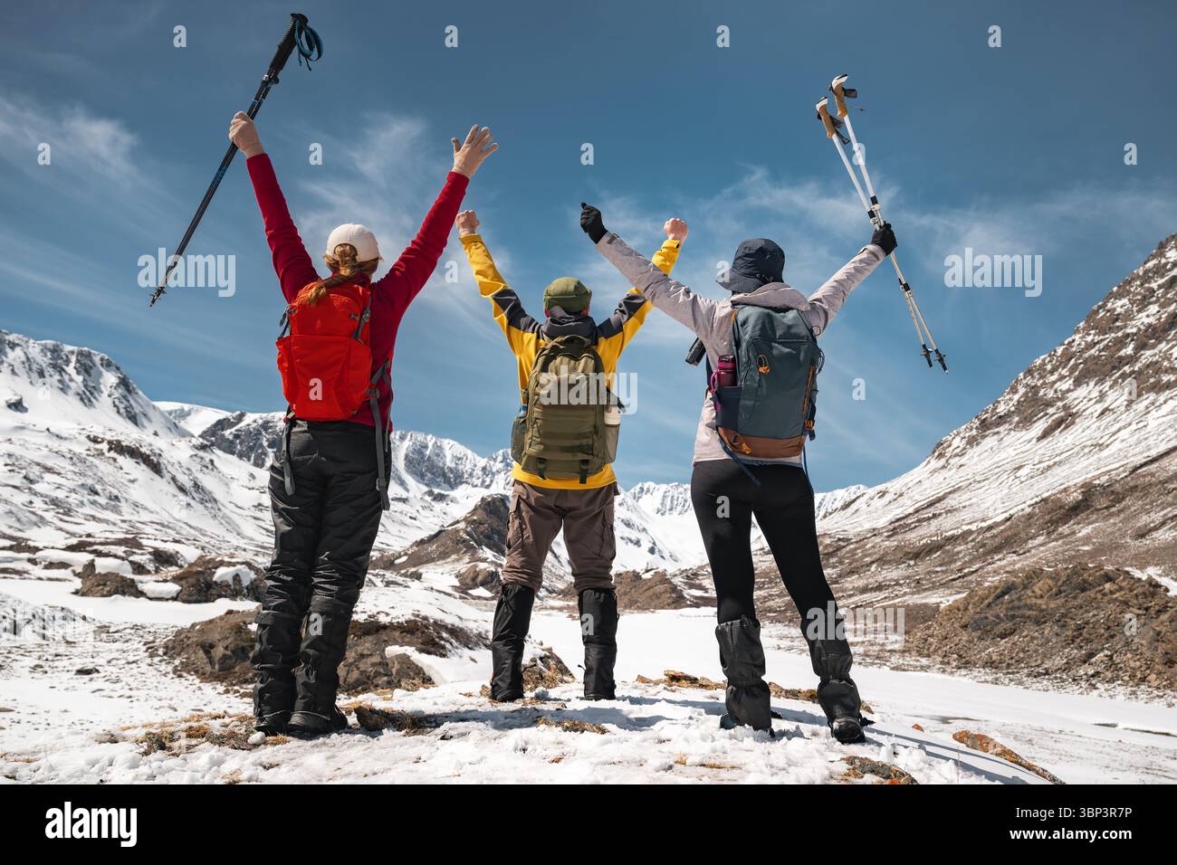 Rückansicht einer Gruppe von drei Wanderern, die mit erhobenen Armen stehen und das Panorama eines verschneiten Bergtals und hoher Gipfel in Altai betrachten. Outdoor-Aktivitäten an sonnigen Wintertagen Stockfoto