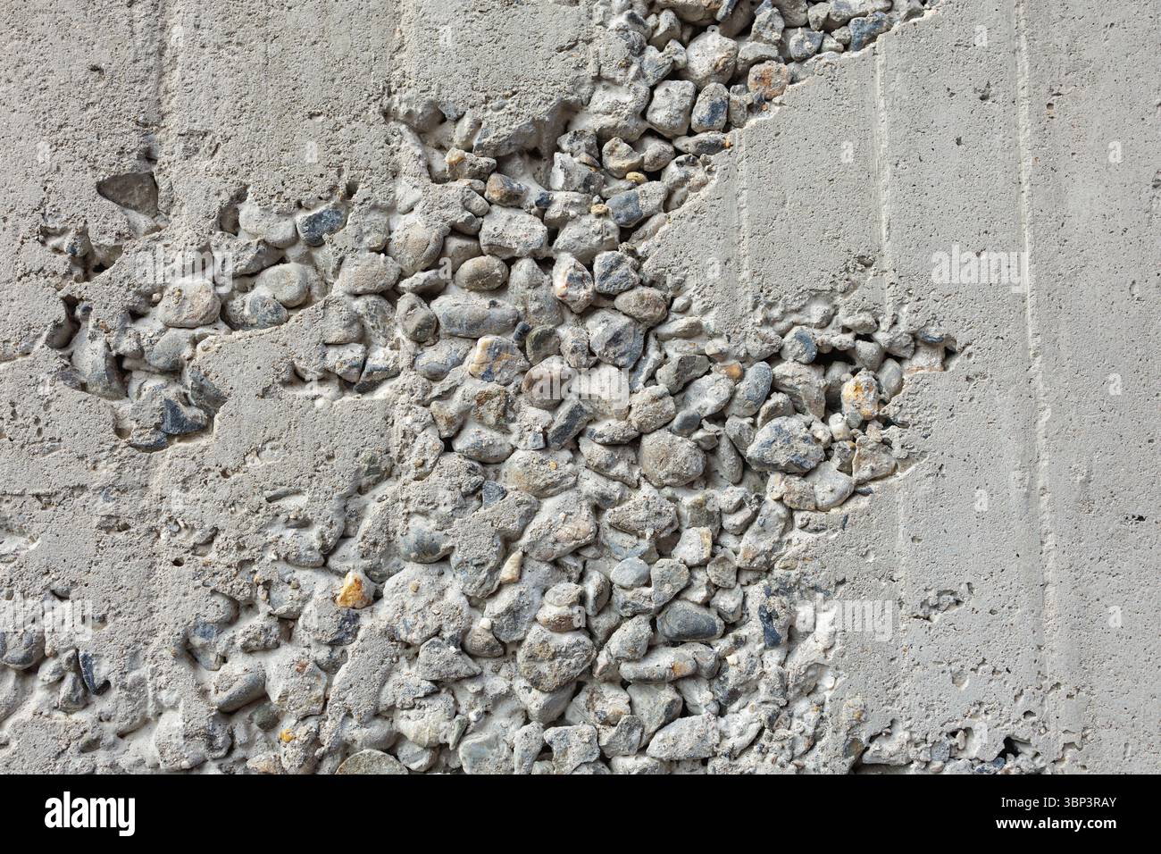 Die raue Betonwand zeigt freiliegende Zuschlagstoffe, die eine robuste Mischsteinoberfläche freigeben. Kontrastierende Texturen heben die eingebettete Struktur von Rohelementen hervor. Stockfoto