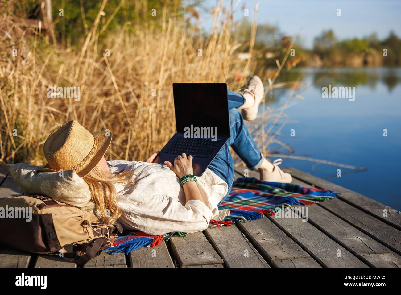Freiberufliche Frau, die sich am See mit einem Laptop zurücklehnt und die Natur genießt, während sie ferngesteuert arbeitet Stockfoto