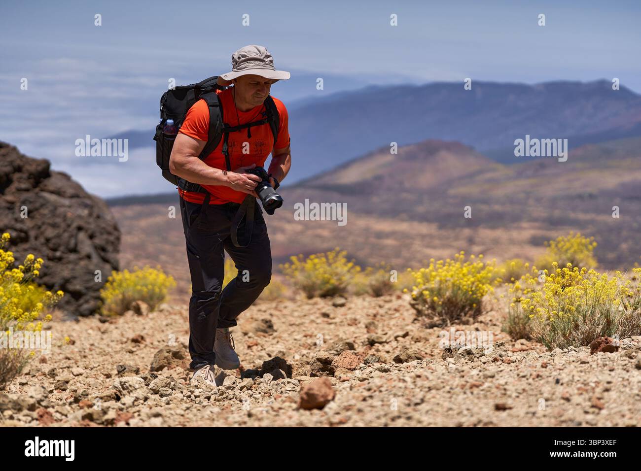 Mann wandert über felsiges vulkanisches Gelände mit Kamera in der Hand und Berglandschaft im Hintergrund Stockfoto