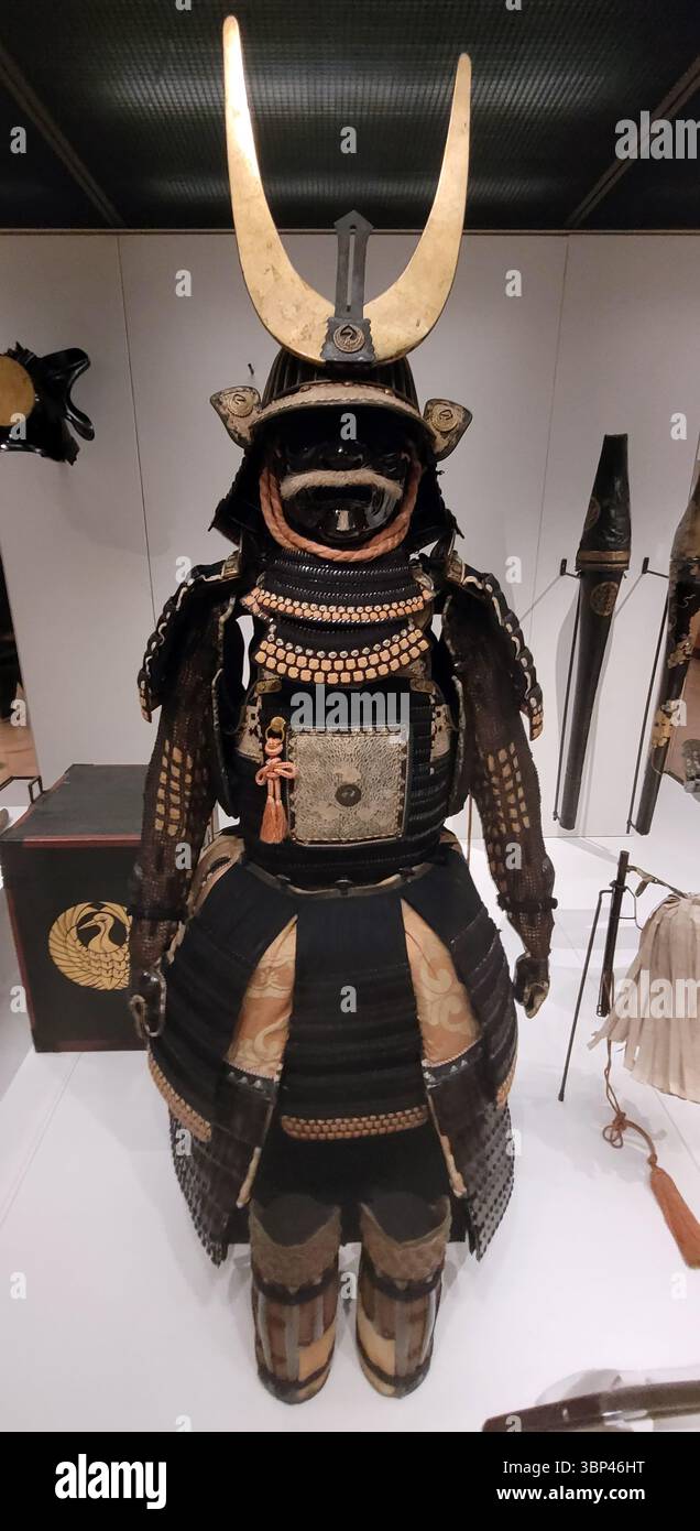 Ein abendfüllendes Foto eines japanischen Samurai-Rüstungsanzugs aus dem 18. Jahrhundert, der zur Familie Mori im Westen Japans gehört, aus Metall, Lack, Leder, Hanffasern, Horn, Holz und Gold. mit großen goldenen Halbmondhörnern und komplizierten Details, ausgestellt in einem Museum. Während der langen Friedenszeit unter dem Tokugawa-Shogunat (Edo-Zeit) verlagerte sich die Funktion der Samurai-Rüstung. Obwohl sie noch in der Lage waren, Schutz zu bieten, wurde der Schwerpunkt zunehmend auf künstlerischem Handwerk, Familienwappen und ausgeklügelten Entwürfen gelegt, die Reichtum und Status zum Ausdruck brachten. Solche Rüstungsstücke werden oft ausgestellt Stockfoto