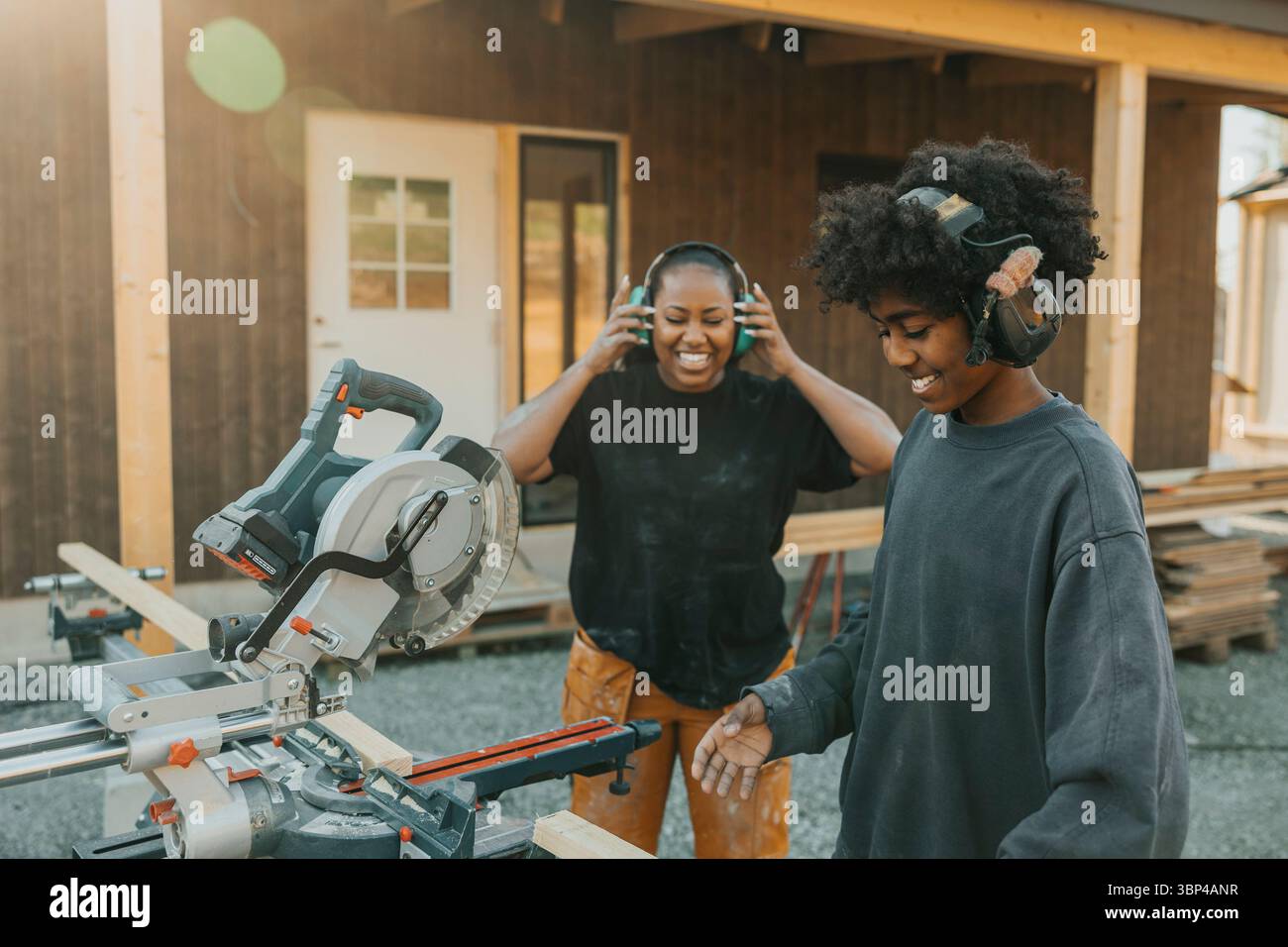Glückliche Mutter, die den Sohn ansieht, der Kopfhörer mit Lärmreduzierung trägt, Holzplanken mit Kreissäge im Hinterhof schneidet Stockfoto