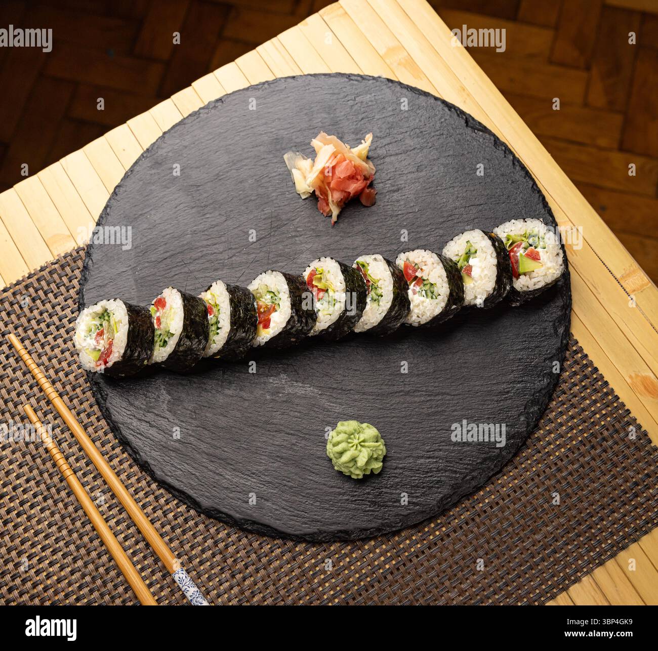 Frisch zubereitete vegane Sushi-Brötchen, gefüllt mit buntem Gemüse, serviert auf einem schwarzen Schieferteller mit Wasabi und eingelegtem Ingwer Stockfoto