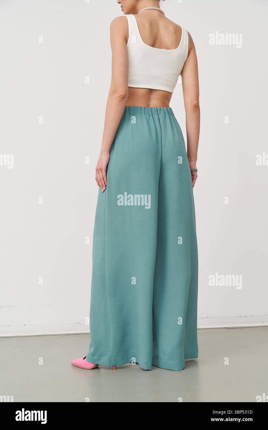 Frau mit hoher Taille, blaublaublauer Hose mit weitem Bein und weißem ärmellosem Crop-Top, seitlich auf hellem Boden. Sie hat pinkfarbene Schuhe und ein B Stockfoto