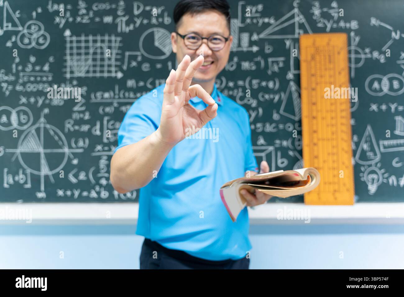 Selbstbewusster, lächelnder Lehrer im Klassenzimmer, der eine gute Handbewegung zeigt, während er das Buch hält, mit mathematischen und naturwissenschaftlichen Formeln vor der Tafel steht, Concept Stockfoto