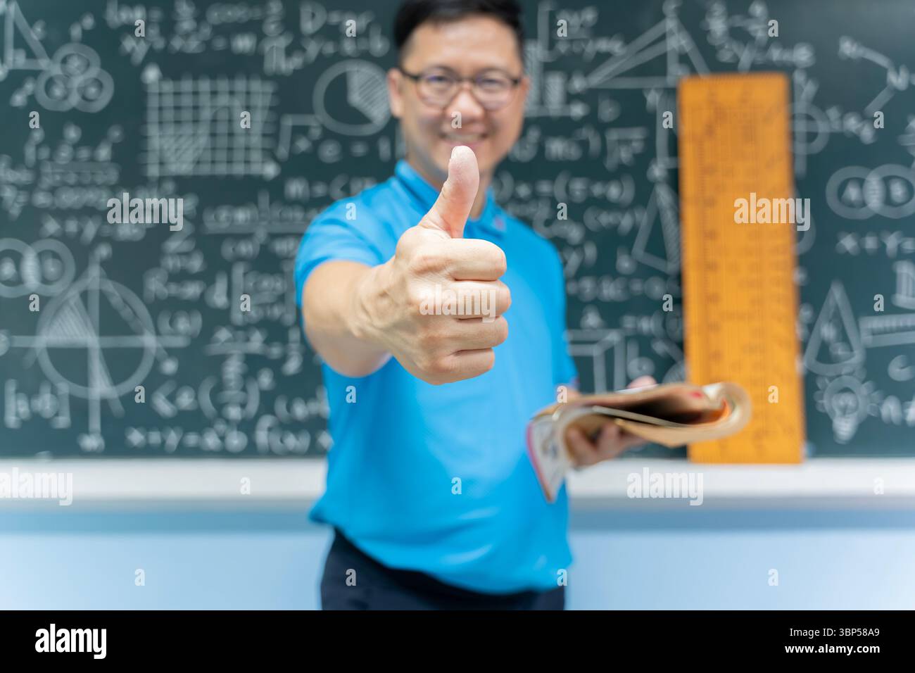 Glücklicher, selbstbewusster Lehrer im Klassenzimmer, der ein Buch hält und die Daumen nach oben zeigt. Er steht mit mathematischen und naturwissenschaftlichen Formeln vor der Tafel Stockfoto