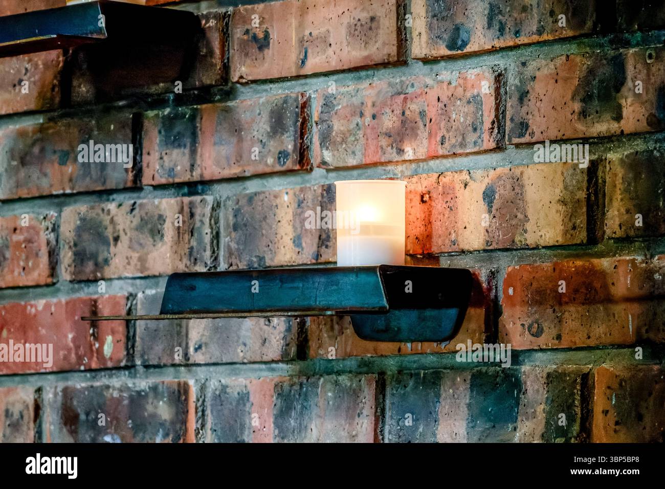 Eine einzelne Votivkerze brennt auf einem Metallregal an einer Ziegelwand in einer Kirche, Symbol für Glauben, Gebet und stille Hingabe Stockfoto