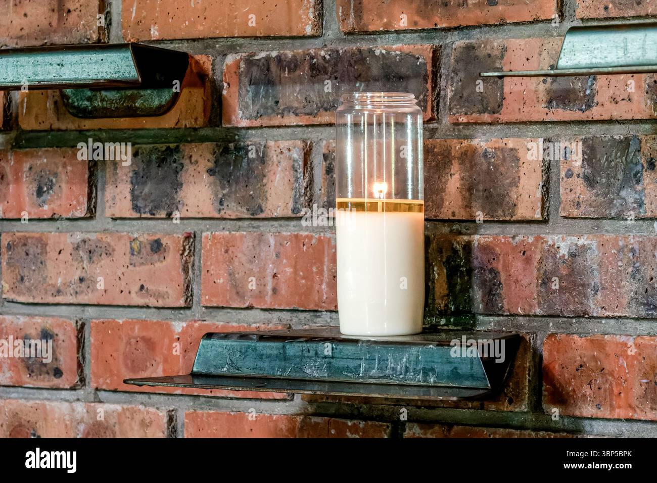 Eine einzelne Votivkerze brennt auf einem Metallregal an einer Ziegelwand in einer Kirche, Symbol für Glauben, Gebet und stille Hingabe Stockfoto