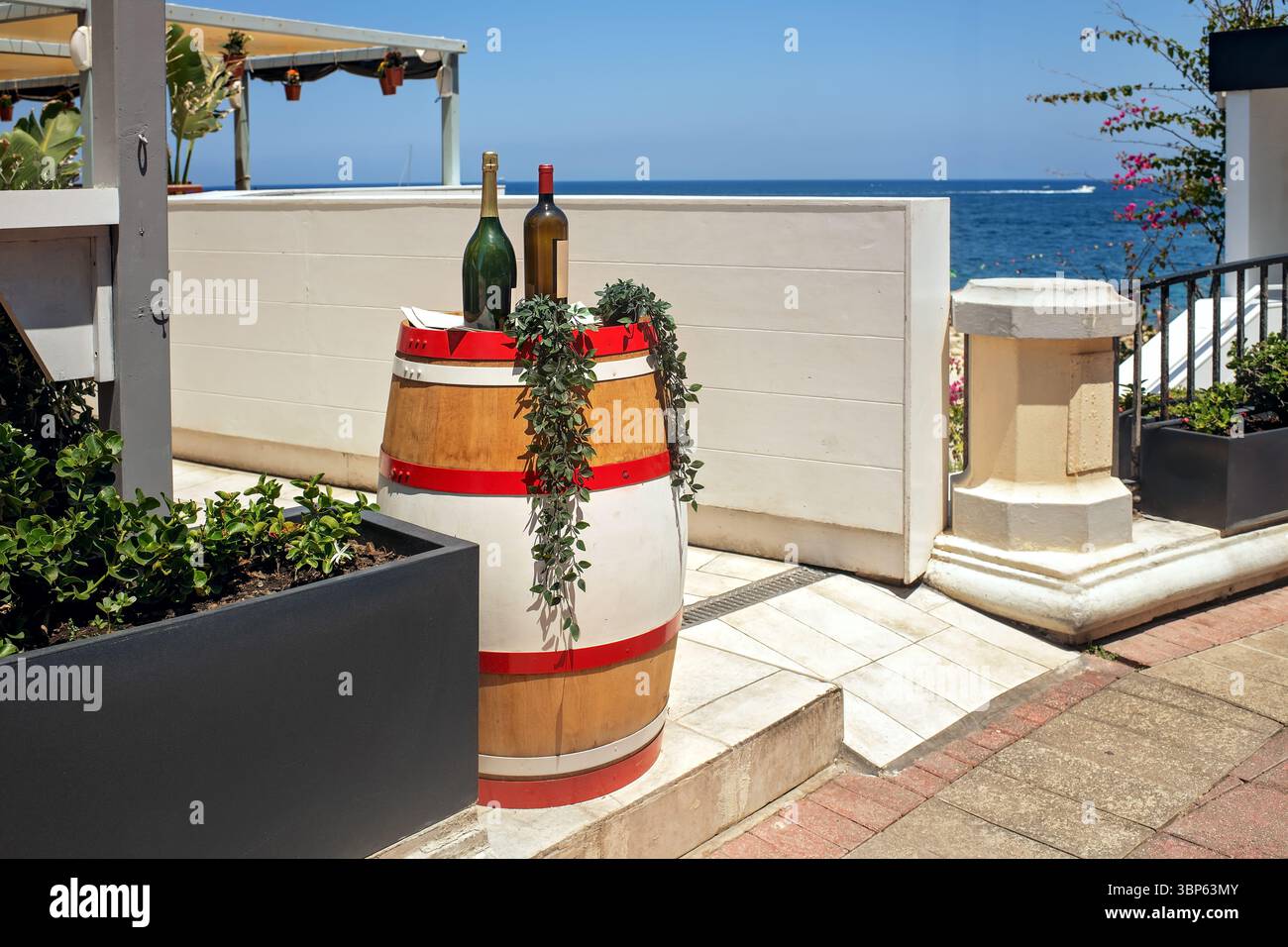 Zwei Glasweinflaschen und Grün auf einem hell bemalten Holzfass auf einer Terrasse am Meer unter blauem Himmel. Konzept von Urlaub, Sommer, Tourismus, und Stockfoto