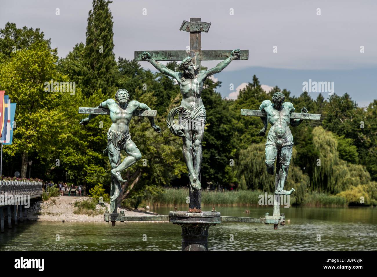 Diese mittelalterliche Bronzeskulptur auf der Brücke zur Insel Mainau in Konstanz zeigt die Kreuzigungsszene mit Jesus Christus in der Mitte und den zwei Dieben an seiner Seite. Die Darstellung ist malerisch über dem Wasser gelegen, umgeben von üppiger Natur Stockfoto