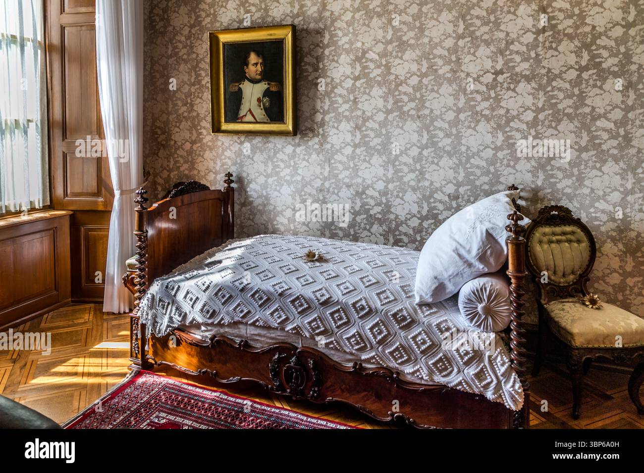 Das historische Schlafzimmer von Louis Napoleon auf Schloss Arenenberg in Salenstein, Thurgau, Schweiz, ist auf dem Bild zu sehen. Das einfache Zimmer ist mit einem antiken Holzbett, einer weißen Bettdecke und einem kunstvollen Stuhl ausgestattet. An der Wand hängt ein Porträt von Napoleon Bonaparte, das die enge Verbindung der Burg zur Familie Bonaparte unterstreicht, da es die ehemalige Residenz von Hortense de Beauharnais war, Napoleons Stieftochter. Das Bild fängt die authentische historische Atmosphäre dieses gut erhaltenen Zimmers ein Stockfoto
