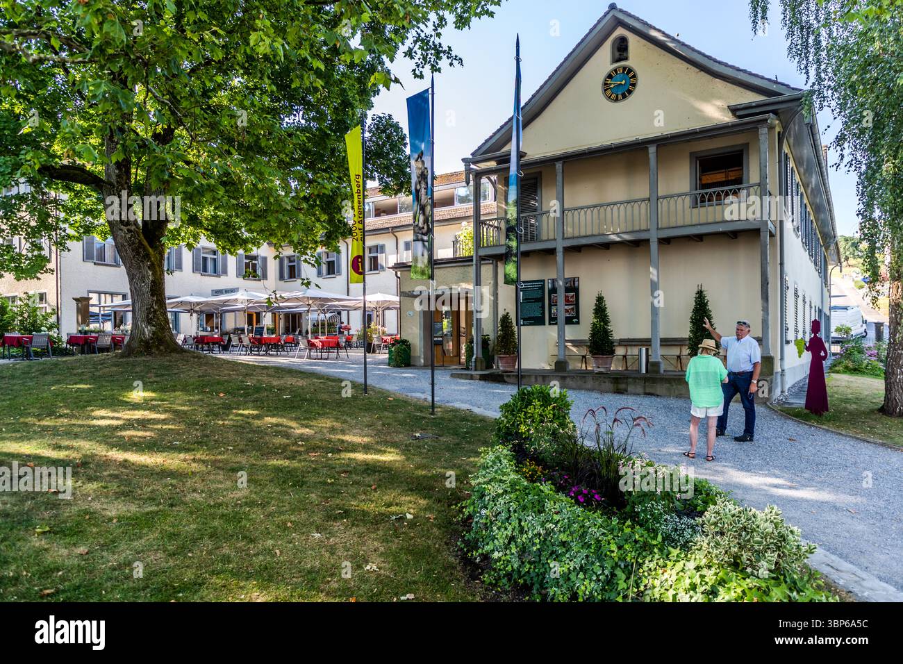 Ein sonniger Tag im Napoleon Museum Arenenberg in Salenstein, Thurgau, Schweiz. Besucher erkunden den Außenbereich des historischen Gebäudes, während andere Gäste die Terrasse des Bistros genießen. Fahnen mit der Aufschrift „Arenenberg“ sind sichtbar Stockfoto