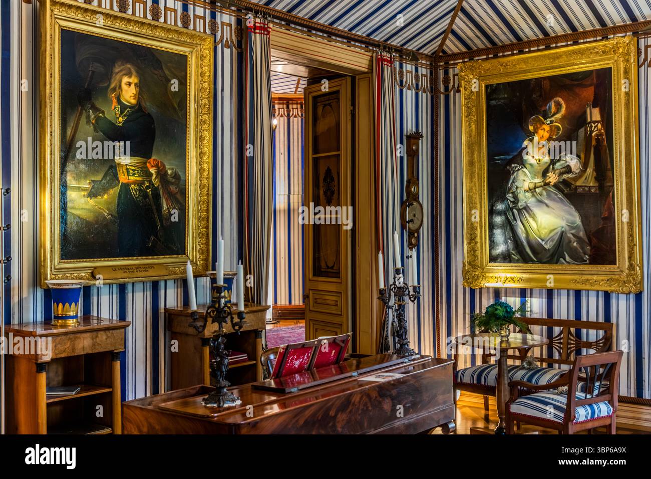 Zeltstube auf Schloss Arenenberg in Salenstein, Thurgau, Schweiz. Das Zimmer ist im Empire-Stil eingerichtet, mit charakteristischen blau-weiß gestreiften Wänden und Decken, die an ein Zelt erinnern. An den Wänden hängen große Porträts, darunter eines von Napoleon Bonaparte und Hortense de Beauharnais. Möbel wie ein Klavier, ein Kerzenleuchter und passende Stühle runden das historische Ambiente ab. Dieser Raum zeugt von der Zeit, als Hortense de Beauharnais, Königin von Holland und Napoleons Stieftochter, hier lebte. Arenenberg, Salenstein, Thurgau, Schweiz Stockfoto