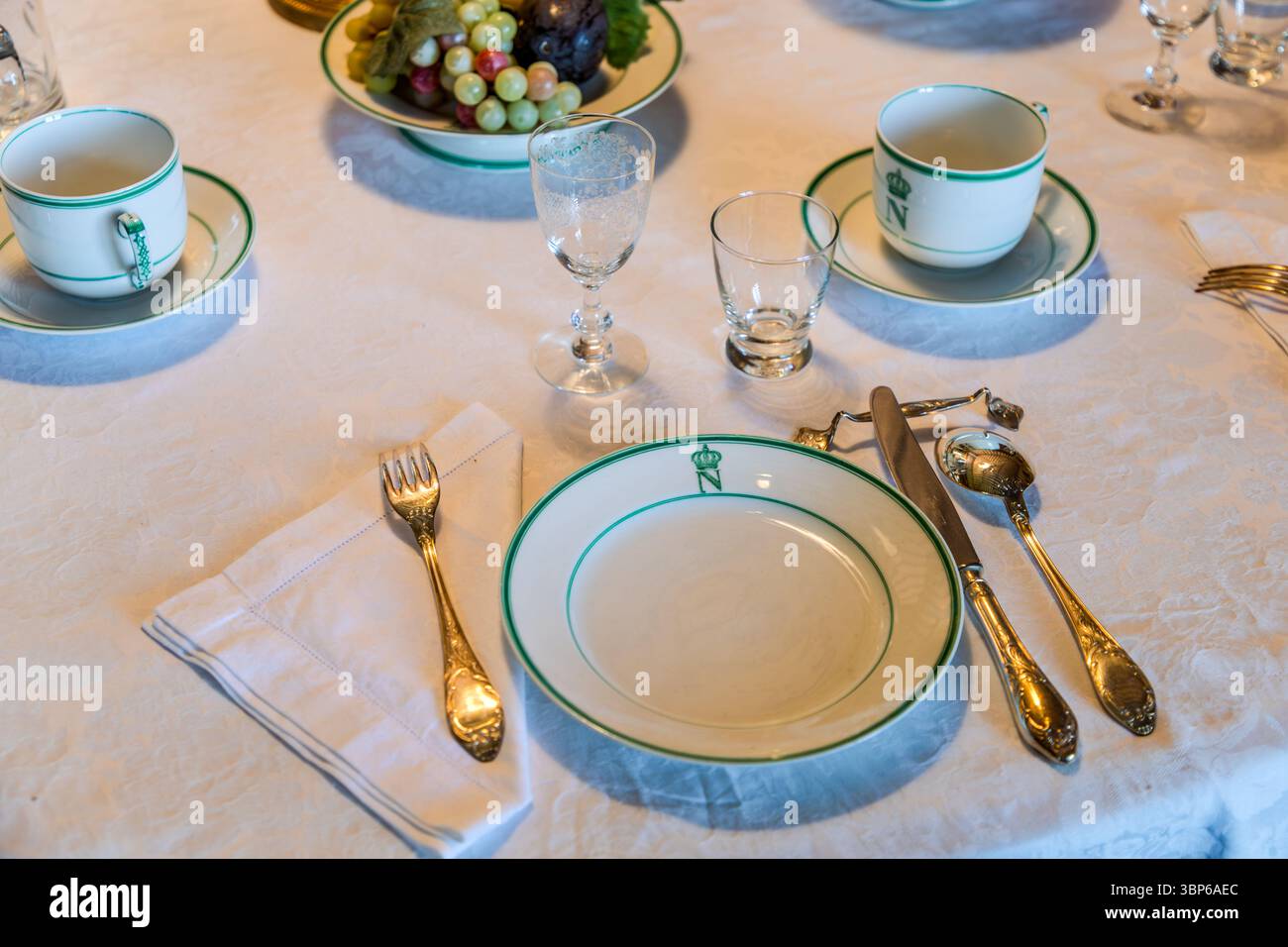 Ein elegant gedeckter Esstisch im Schloss Arenenberg in Salenstein, Thurgau, Schweiz. Das Porzellan von Limoge ist mit einem gekrönten N-Monogramm verziert, das auf die Familie Bonaparte verweist, die hier wohnte. Goldfarbenes Besteck, feine Gläser und eine Obstschale runden das historische Ambiente eines festlichen Tisches ab Stockfoto