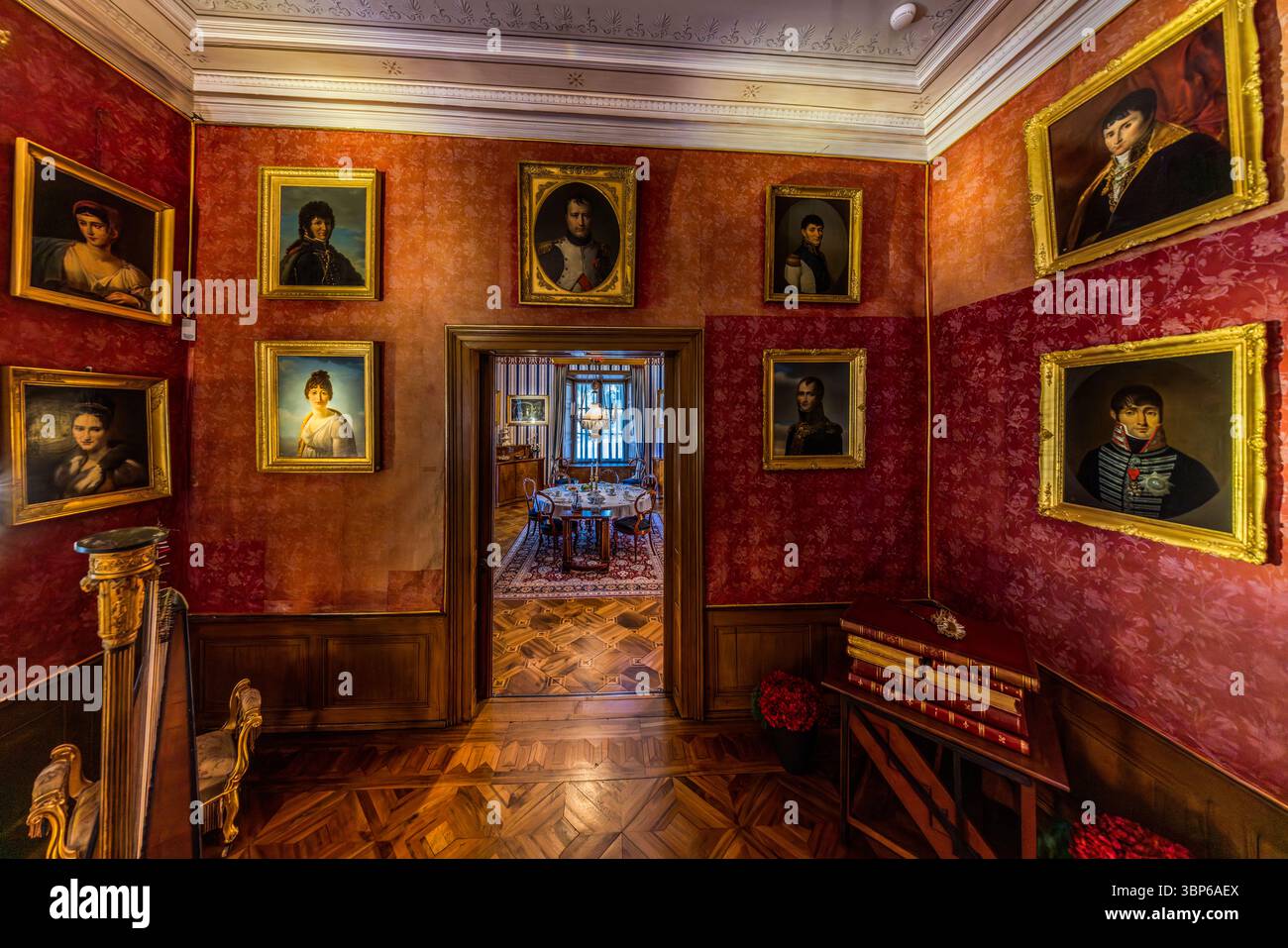 Der Raum im Napoleon-Museum mit roten, gemusterten Wänden zeigt Porträts von Mitgliedern der Familie Bonaparte. Eine Harfe steht im Vordergrund links. Auf der rechten Seite steht ein Stapel Bücher auf einem Tisch. Eine offene Tür führt in ein Nachbarzimmer mit Esstisch. Der Boden besteht aus Holz mit einem eingelegten Muster. Der Standort ist Arenenberg in Salenstein, Thurgau, Schweiz Stockfoto