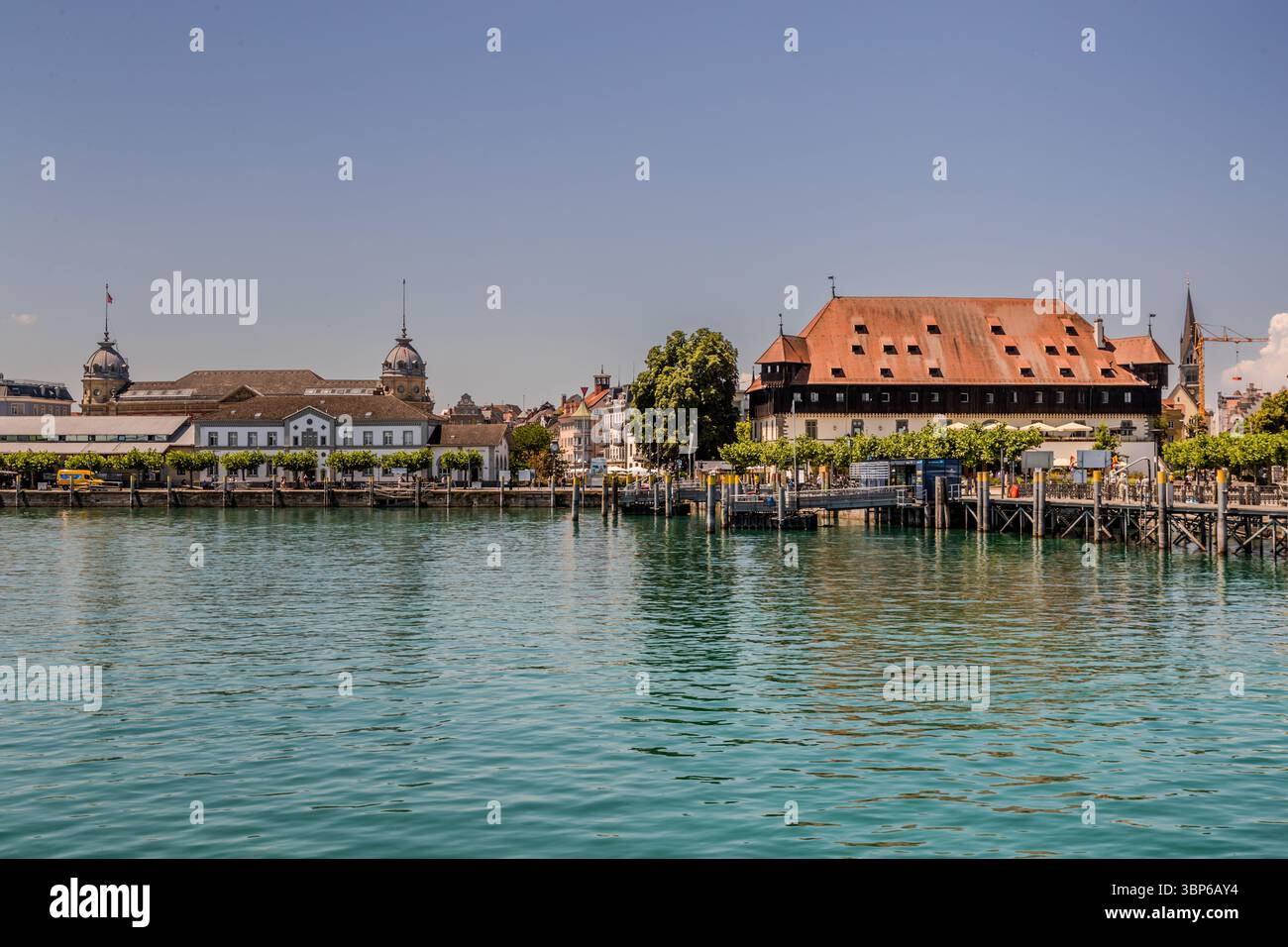 Konstanzer Hafen am Bodensee. Das Konzil-Gebäude und der Hafenbahnhof säumen das Ufer. Die Leute sind auf der Promenade. Stadtgarten, Konstanz, Baden-Württemberg, Deutschland Stockfoto