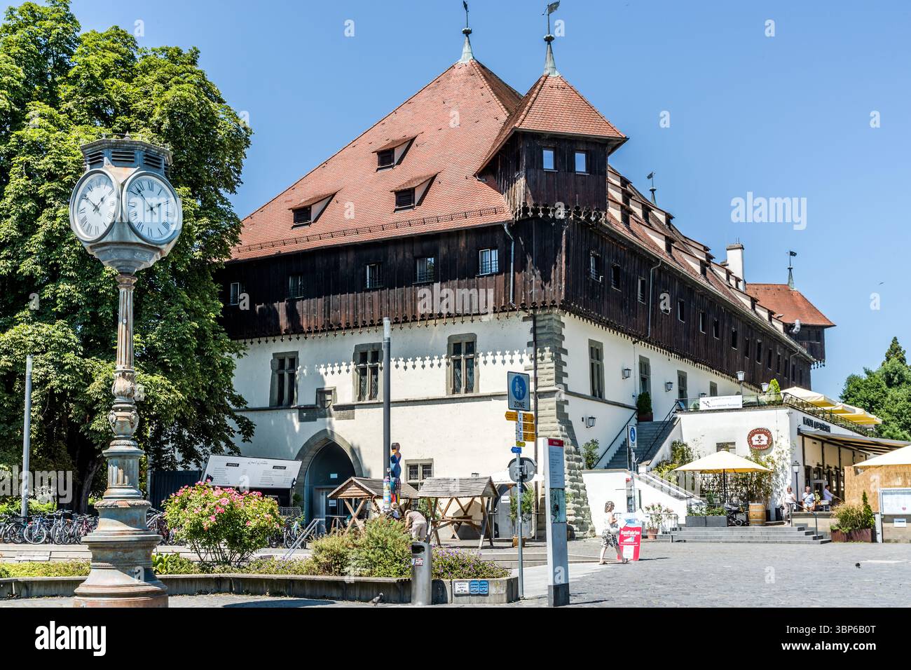 Das Konzilgebäude befindet sich an der Hafenstraße in Konstanz, Baden-Württemberg. Im Vordergrund ist ein Uhrmast zu sehen. Die Leute laufen und sitzen auf dem Platz Stockfoto