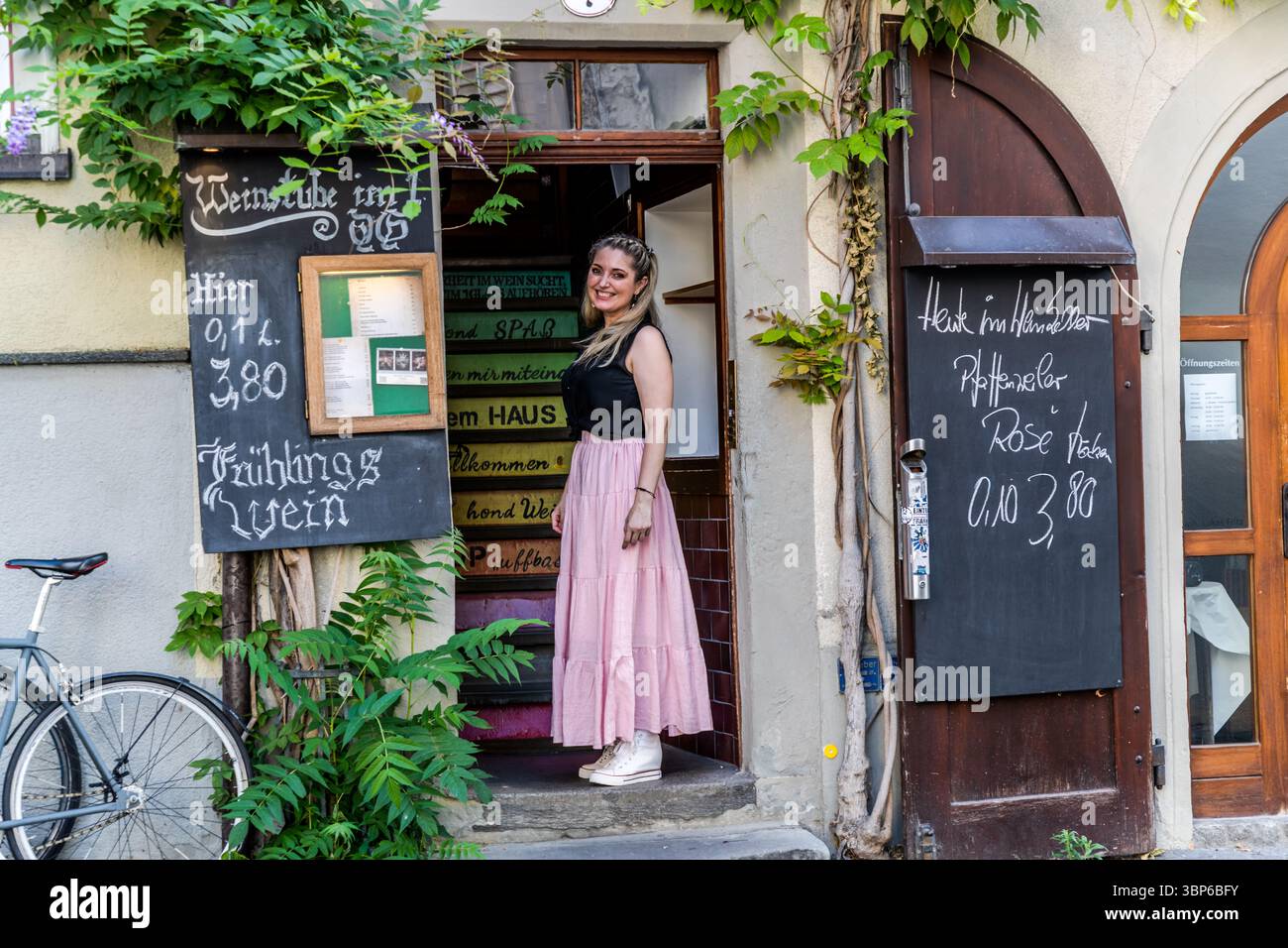 Die Wirtin steht lächelnd am Eingang einer Weinbar in Konradigasse, Konstanz. Vor dem Gebäude befinden sich Tafeln mit Sonderangeboten und ein Fahrrad. Konradigasse, Konstanz, Baden-Württemberg, Deutschland Stockfoto