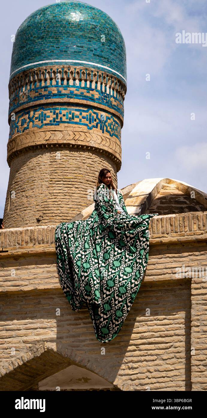 Eine Frau in einem fließenden grün-weiß gemusterten Kleid sitzt anmutig auf einem historischen Gebäude mit einer blauen Kuppel. Die Architektur zeichnet sich durch eine komplizierte Gestaltung aus Stockfoto