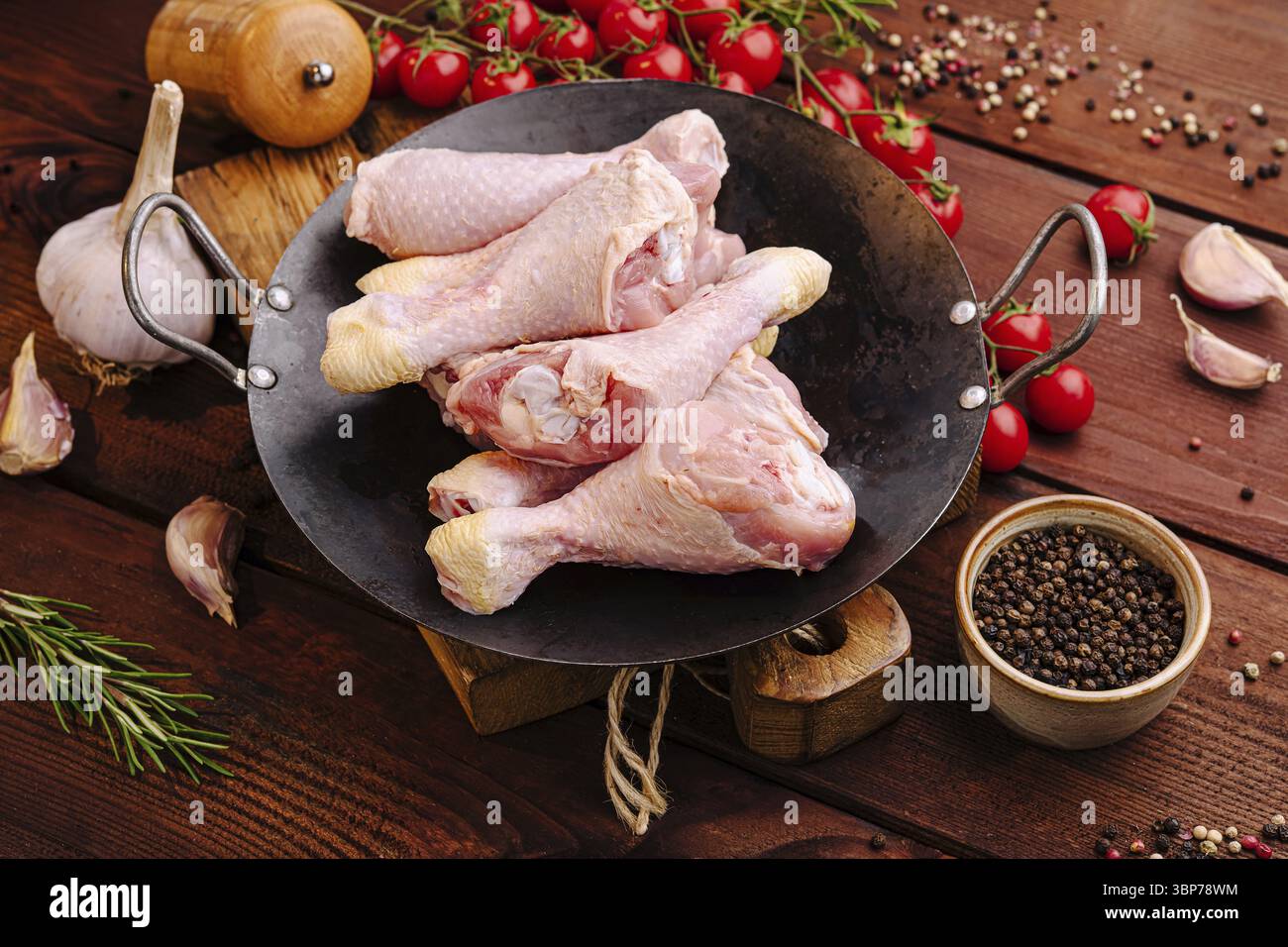 Speisen, Eine Auswahl an rohen Hähnchenstangen wird in einer schwarzen Pfanne neben Kirschtomaten und Gewürzen auf einem Holztisch präsentiert, auf dem eine Invi präsentiert wird Stockfoto