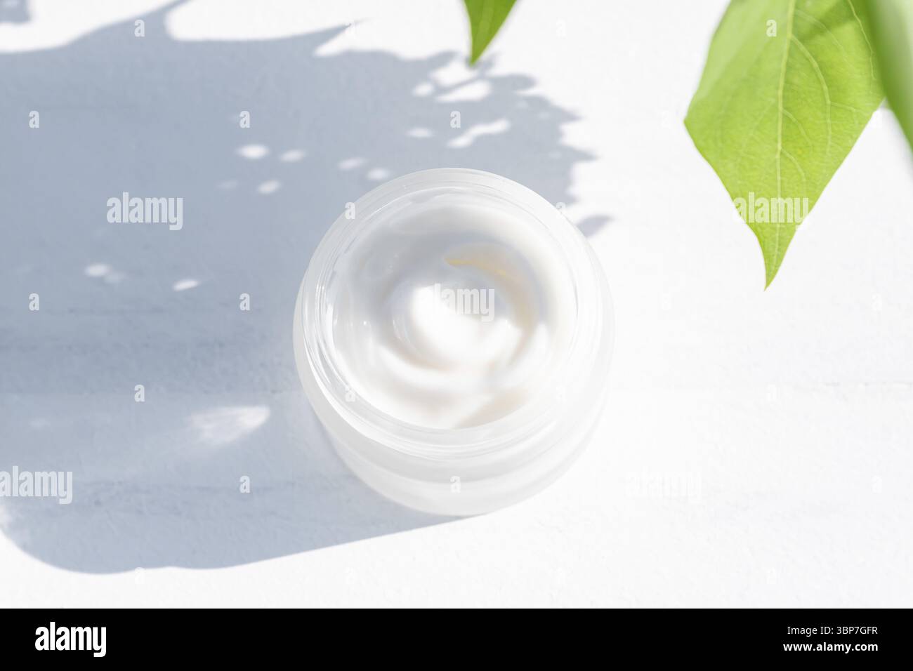 Nahaufnahme von Kollagencreme im Glasgefäß und grünen Blättern auf weißem hölzernem Hintergrund. Schroffe Schatten. Kosmetikprodukte für Haut- und Körperpflege Stockfoto