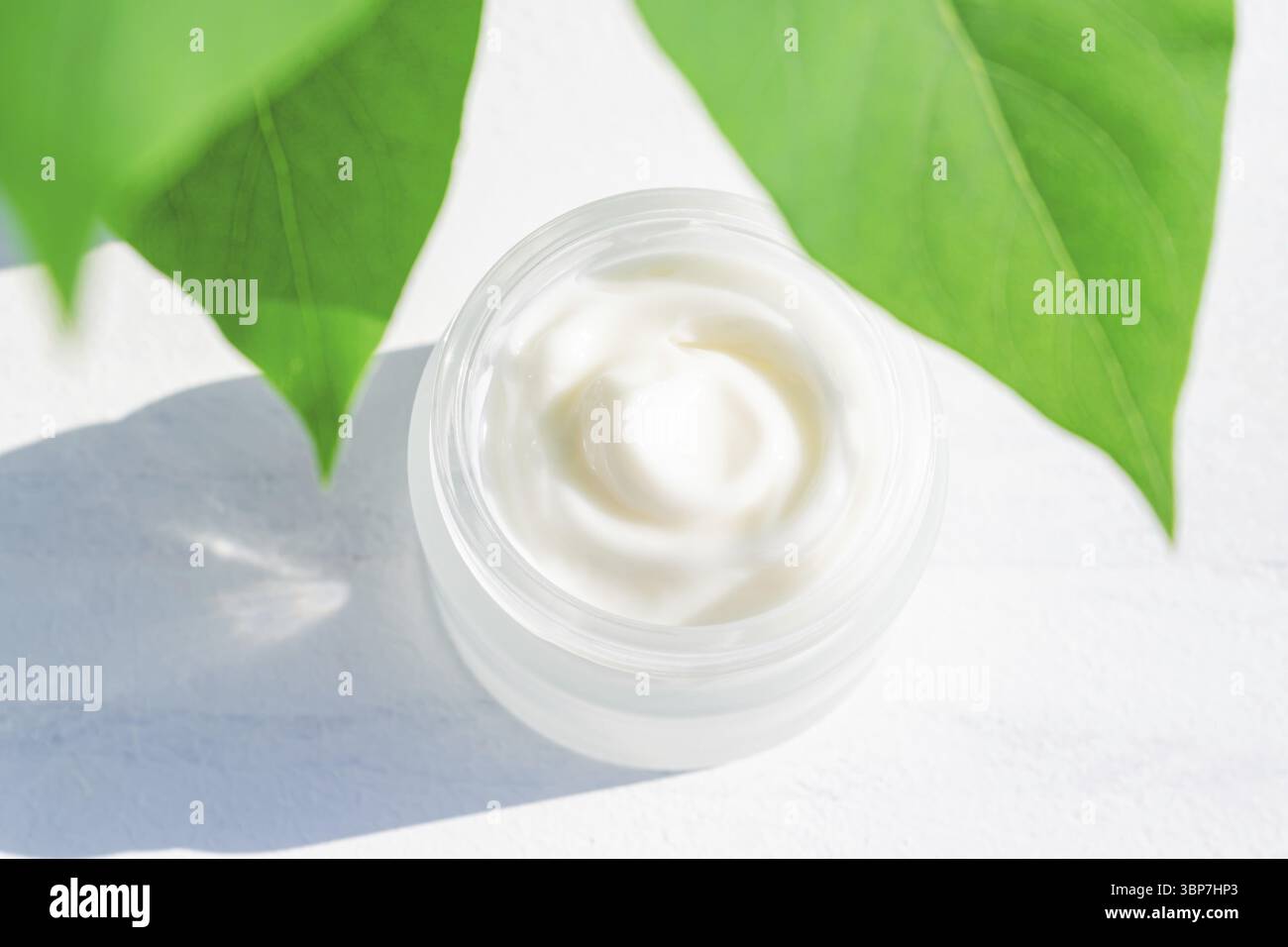 Feuchtigkeitscreme-Glas in Milchglasflasche und grünen Blättern auf weißem Hintergrund. Nahaufnahme der Textur von Haut- und Körperpflegeprodukten Stockfoto