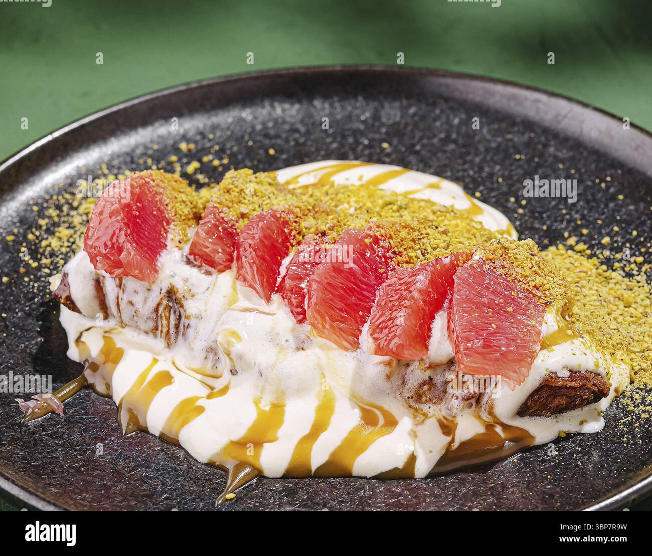Food, Ein köstliches Dessert, besteht aus cremigen Joghurtschichten mit frischen pinken Grapefruitscheiben und zerstoßenen Pistazien auf einem dunklen Teller. Das Gericht Stockfoto