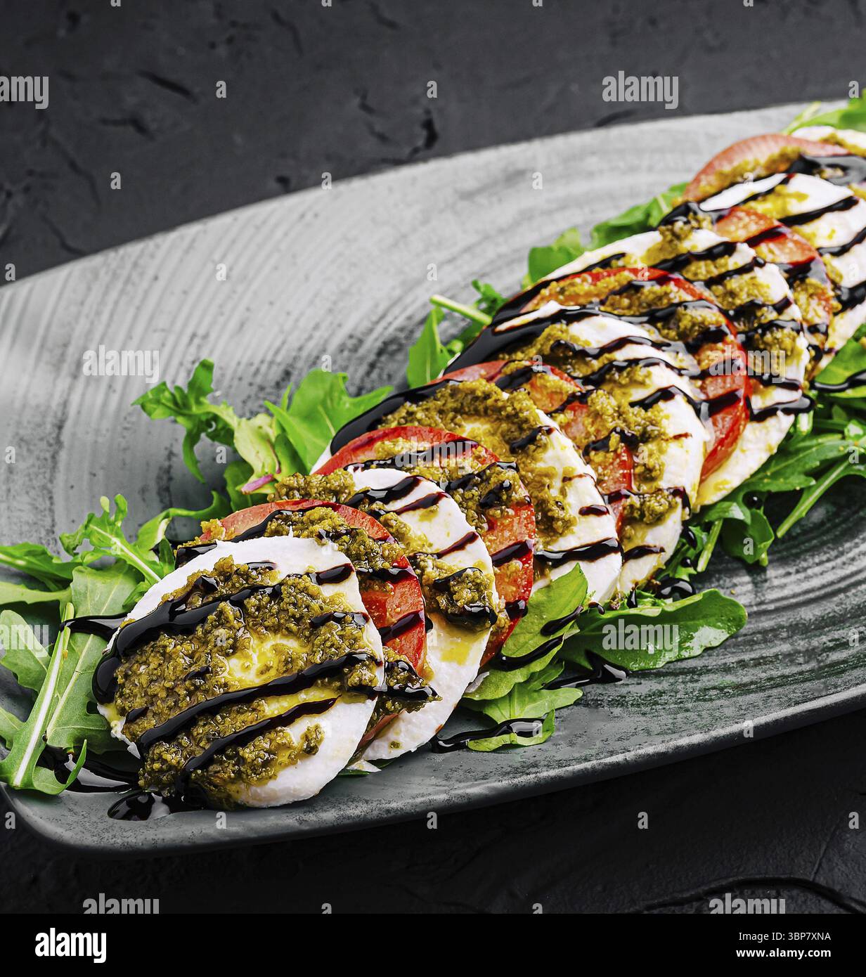 Essen, frische Mozzarella-Scheiben, mit Reifen Tomaten und Pesto, garniert mit Rucola und einem balsamischen Niesel, elegant auf einem dunklen Servo präsentiert Stockfoto